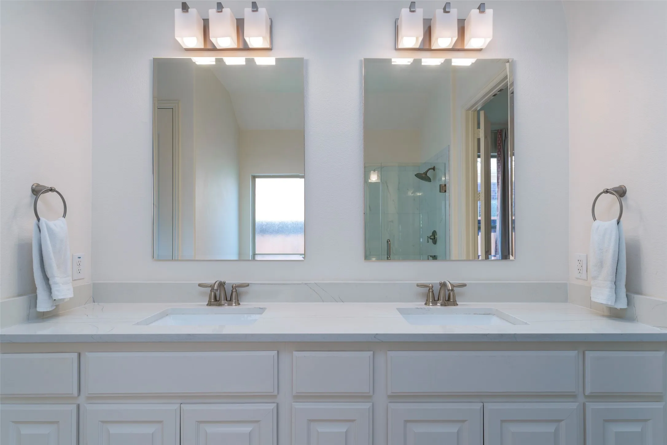 Updated primary bathroom vanity featuring dual sinks, generous quartz countertop space, and crisp white cabinetry. Frameless mirrors, modern light fixtures, and updated plumbing fixtures add a fresh, contemporary touch to this bright and functional space.