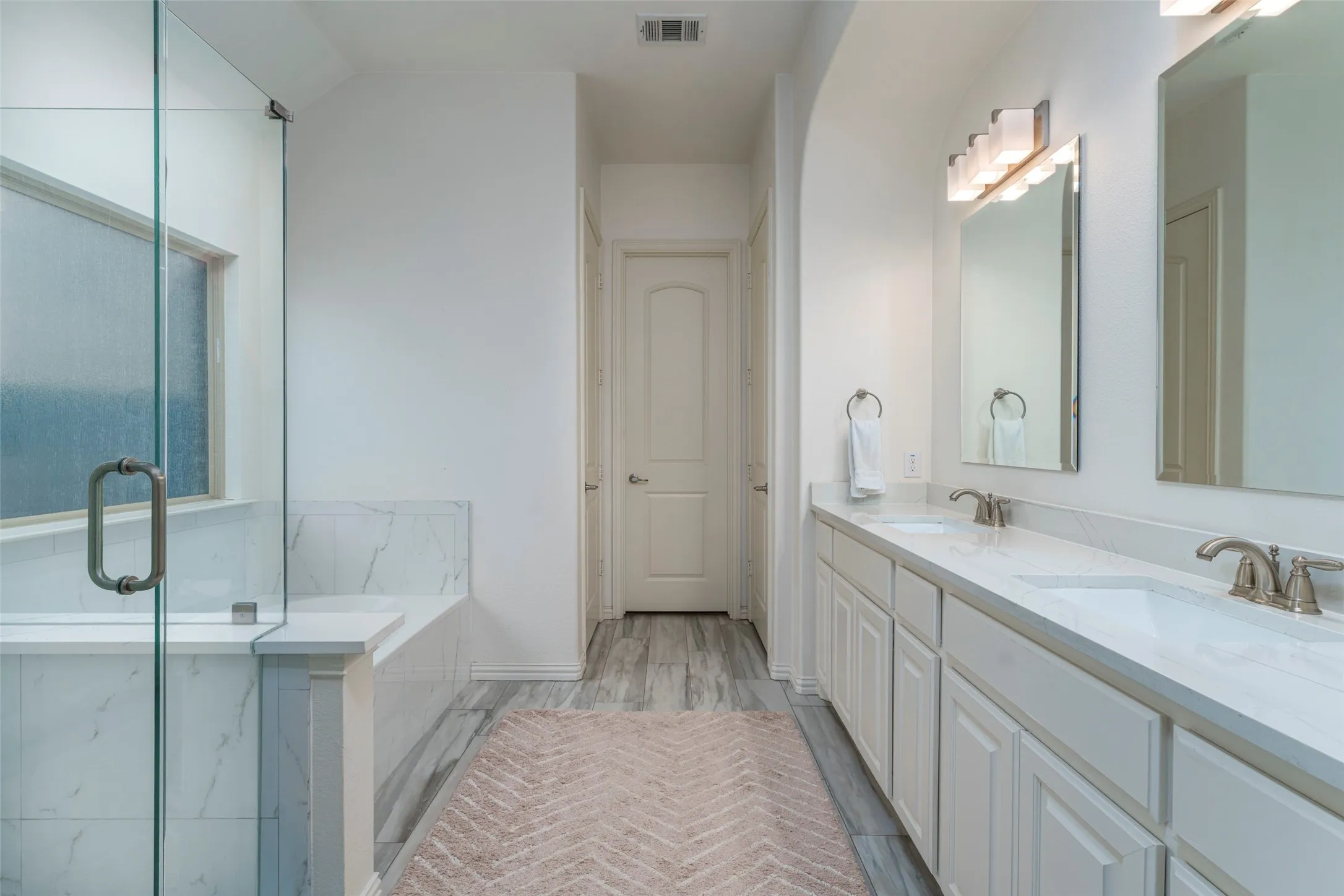 Spa-inspired primary bathroom featuring taller white vanities with updated quartz countertops and dual sinks. A seamless glass-enclosed shower and separate soaking tub create a relaxing retreat, complemented by neutral tile flooring and a bright, timeless design.