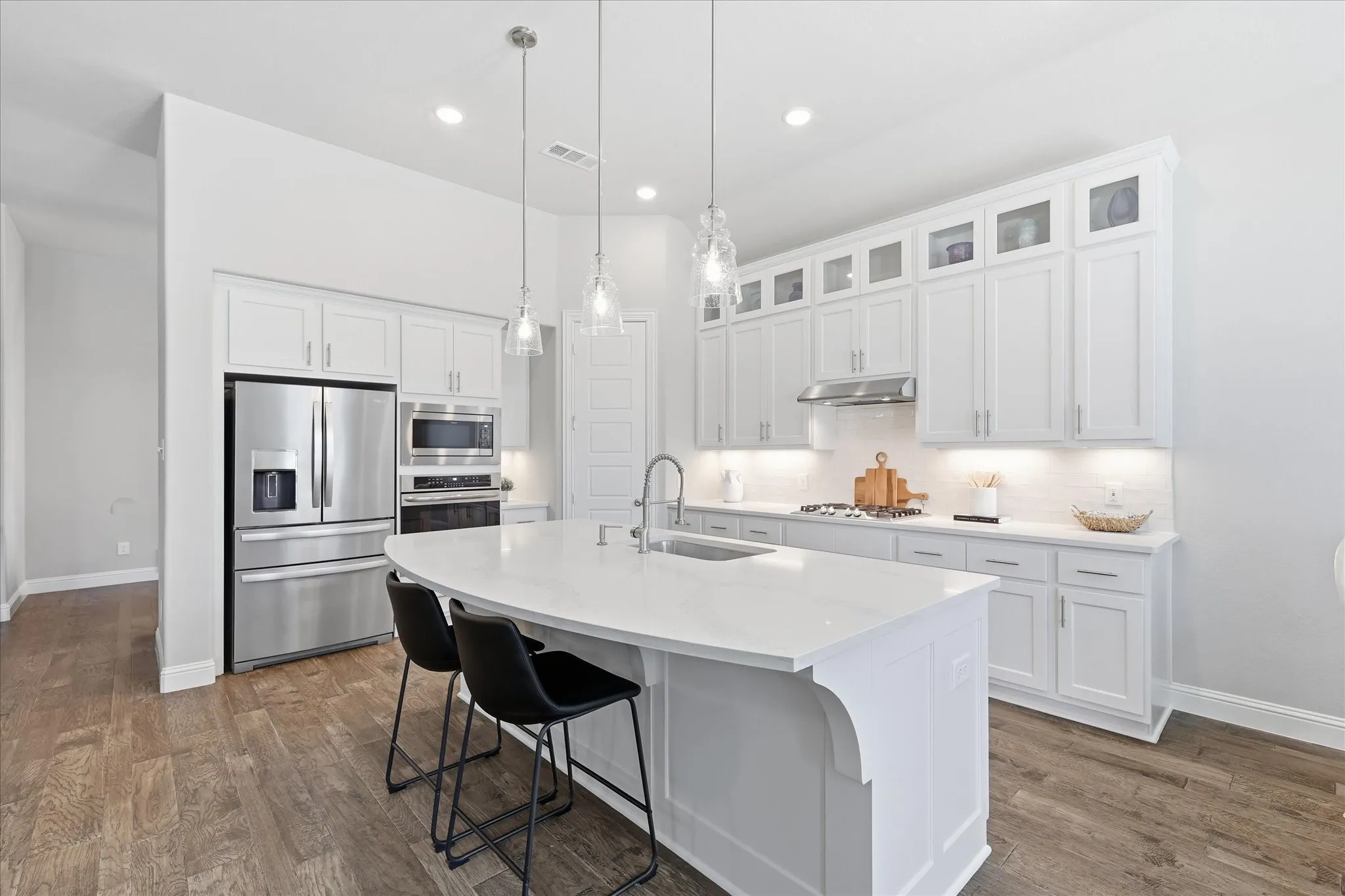 Expansive kitchen island with a white countertop, stainless steel appliances, white shaker cabinets, wood-finish flooring, and pendant lighting