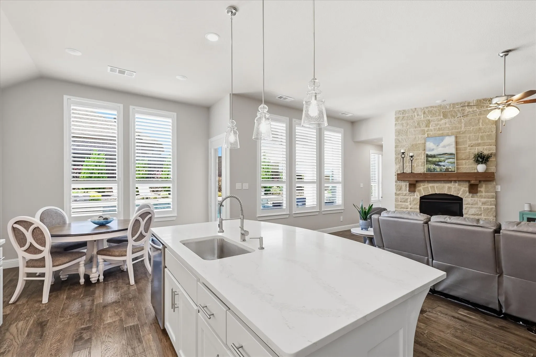 Spacious kitchen island with a built-in sink, stone-clad fireplace with a wood mantel, wood-finish flooring, recessed lighting, and multiple windows with blinds