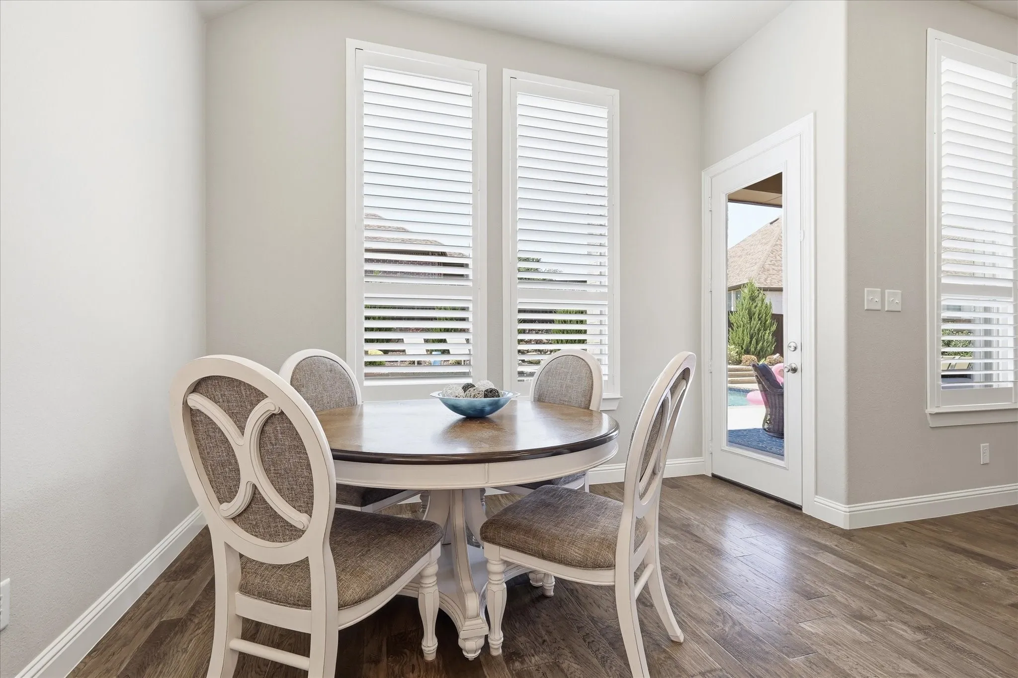 Wood-finish flooring with white baseboards, bright white wall paint, multiple windows with white plantation shutters, and a glass-paneled door