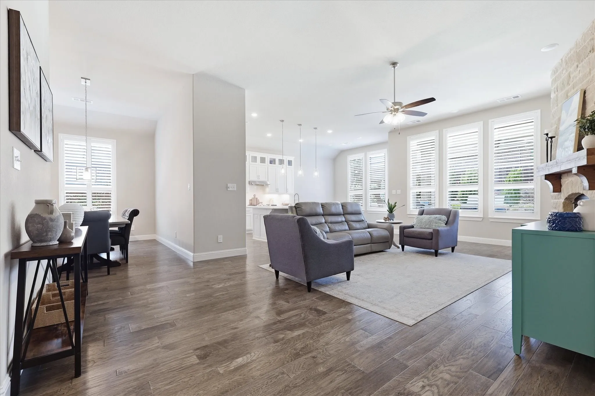 Wood-finish flooring throughout, white plantation shutters, stone accent wall with wood mantle, ceiling fan, and recessed lighting