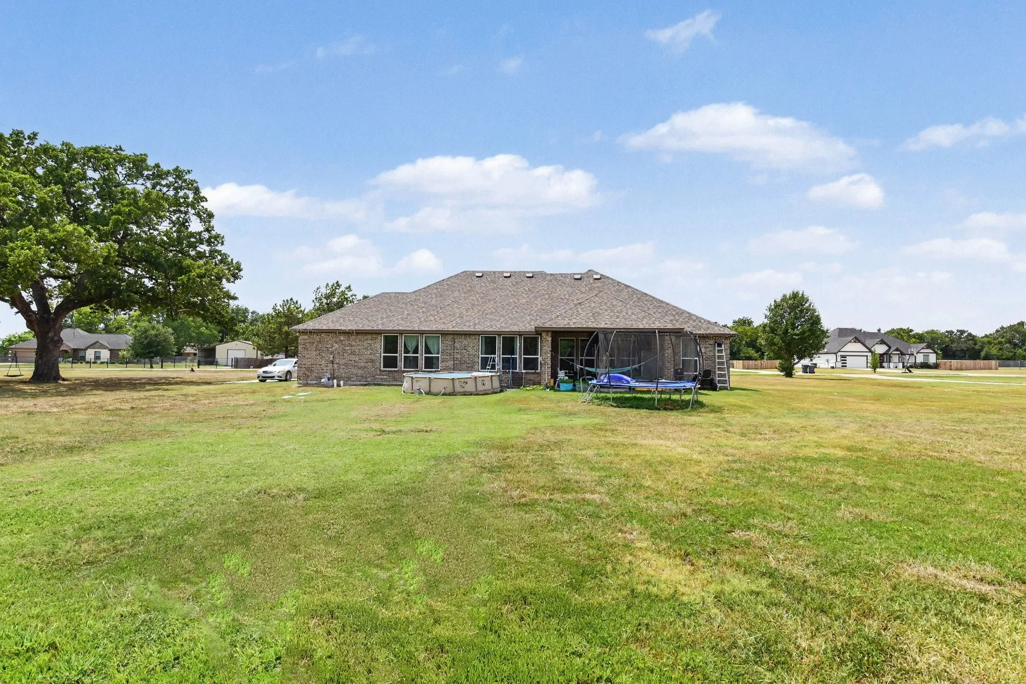 Expansive grassy yard with mature trees, brick exterior, multiple windows, and a covered patio