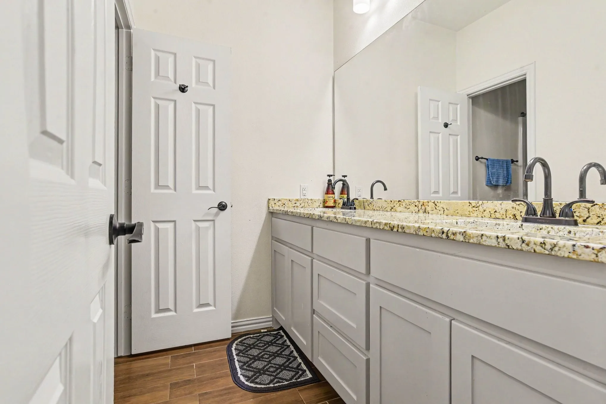 Dual sink vanity with granite countertop, gray cabinetry, dark fixtures, and wood-finish flooring