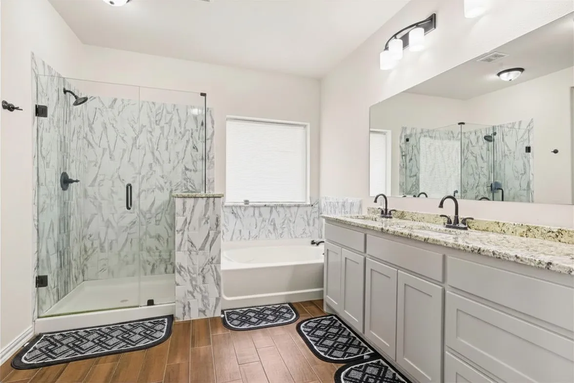 Walk-in shower with clear glass enclosure, a built-in tub, dual vanity with undermount sinks, wood-look ceramic tile flooring, and a large wall-mounted mirror