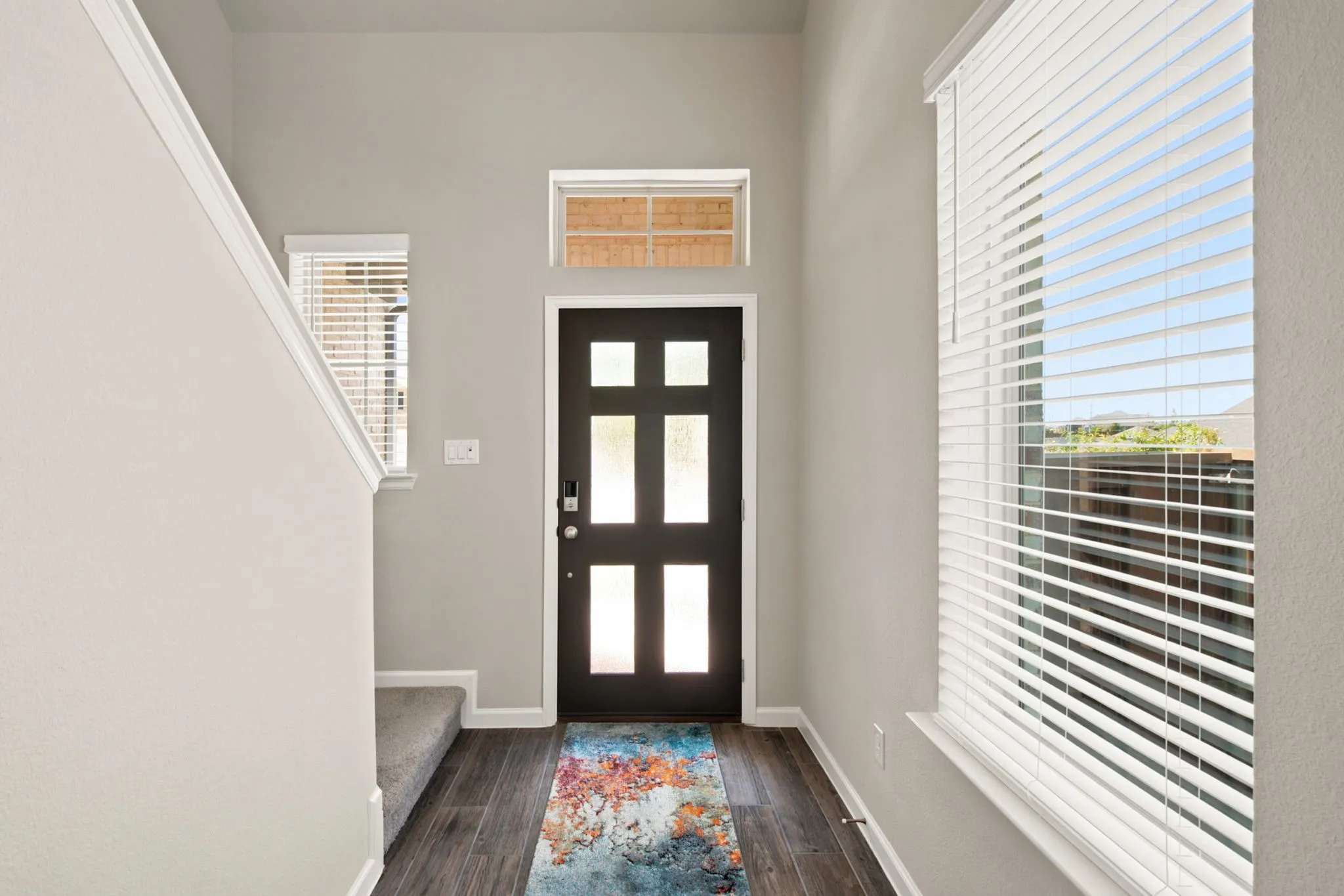 Welcoming entryway showcasing a six-panel black interior door with frosted glass panes, dark wood-finish flooring, and bright natural light from expansive windows with horizontal blinds