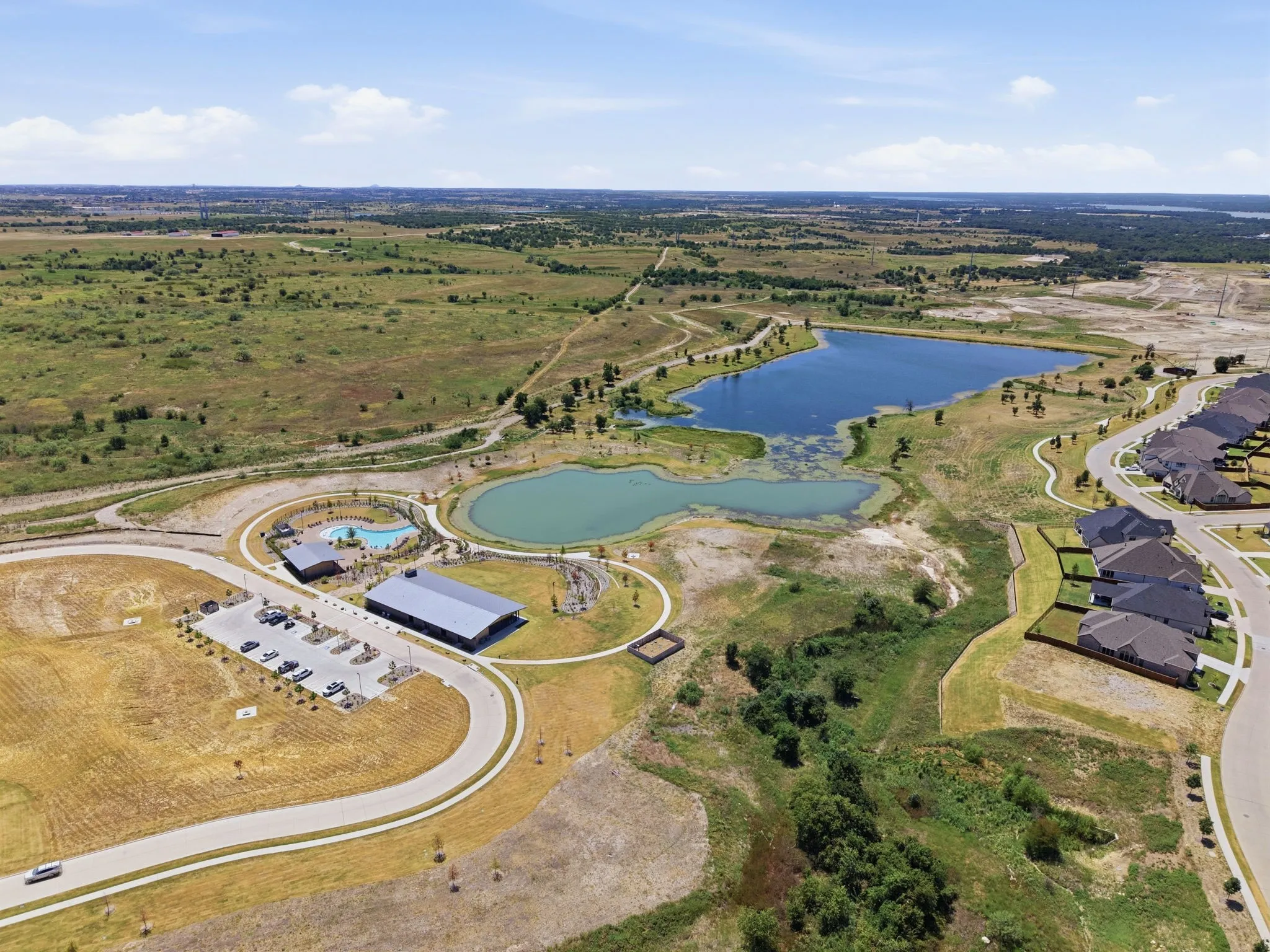 Aerial perspective of a developing community featuring a large lake, a second smaller pond, a community pool, a large covered amenity structure, and paved residential streets with newly constructed homes