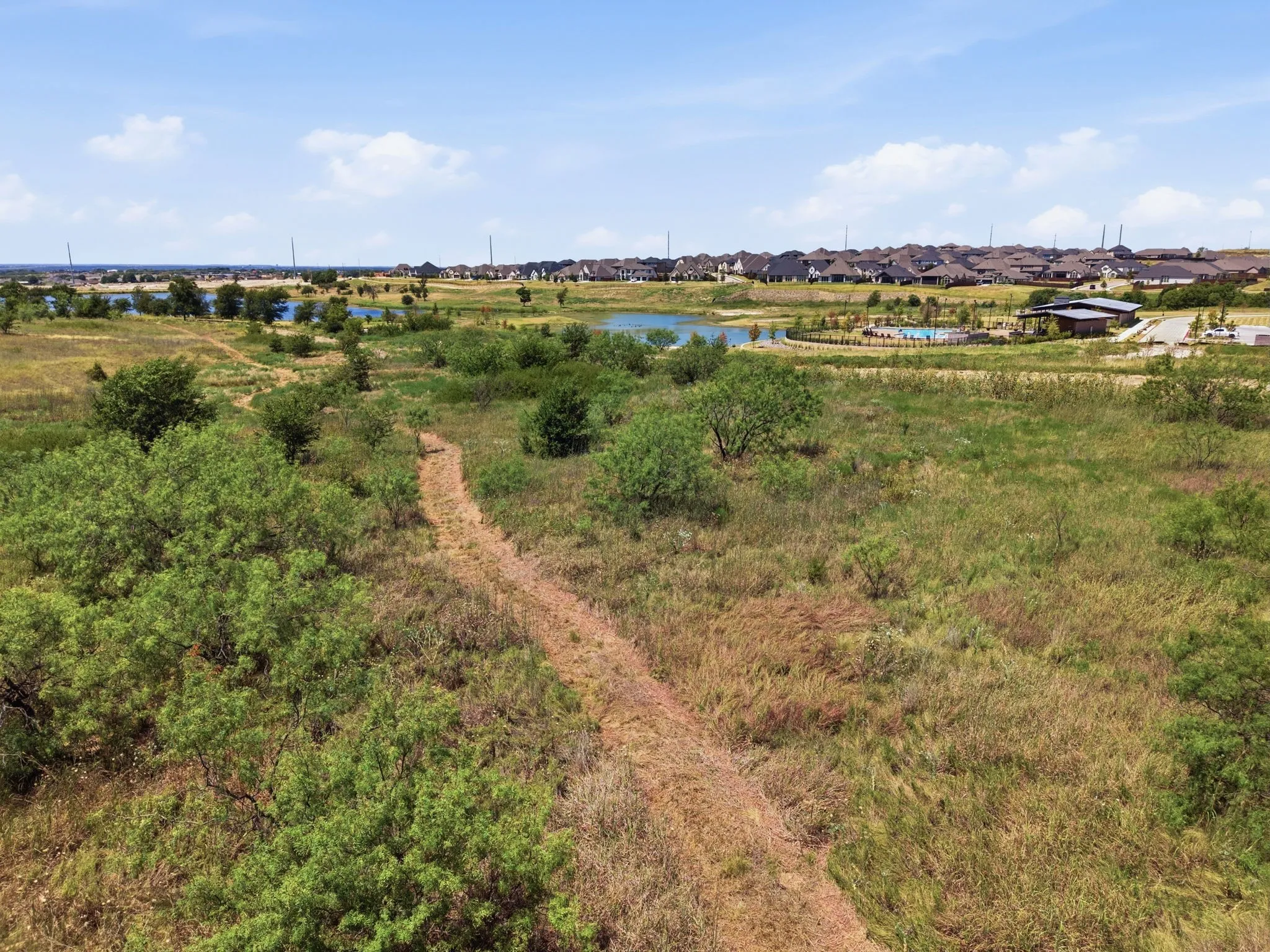Expansive natural landscape with established vegetation, a visible dirt path, a body of water, and residential structures in the distance