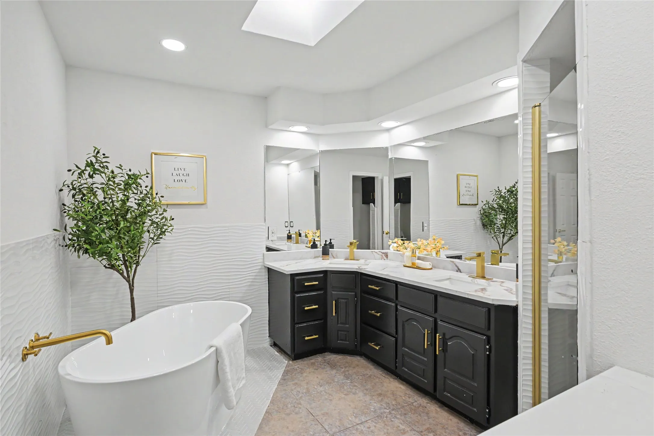 Spa-inspired primary bathroom featuring a freestanding soaking tub with brass fixtures, dual-sink vanity with black cabinetry and brass hardware, white subway tile wall treatment, quartz countertops, large framed mirror, skylight, recessed and pendant lighting, and decorative potted greenery