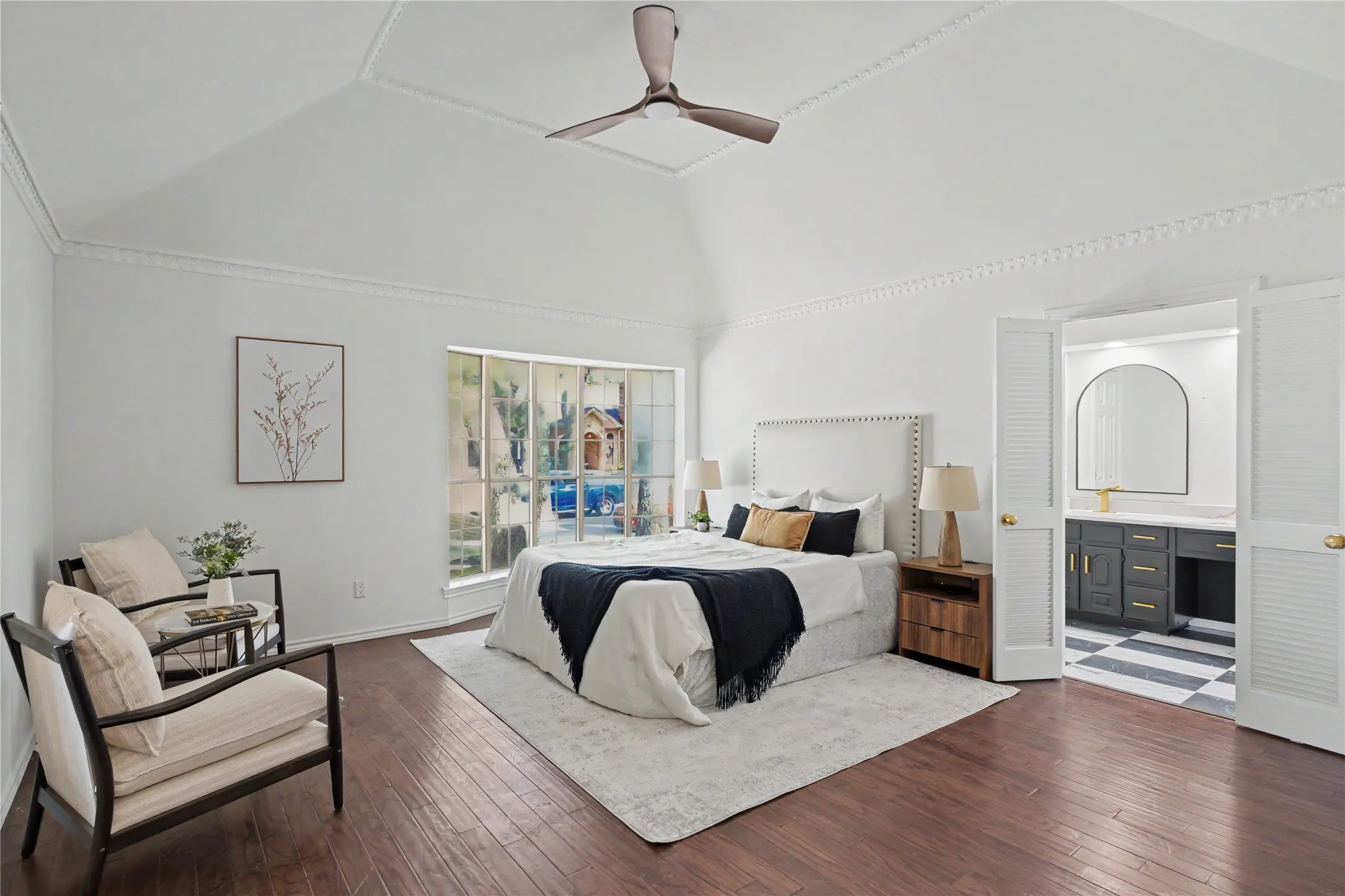 Vaulted ceiling with decorative molding, wood-finish flooring, large multi-pane window, louvered bi-fold doors, and a bathroom with checkered tile and a dual vanity