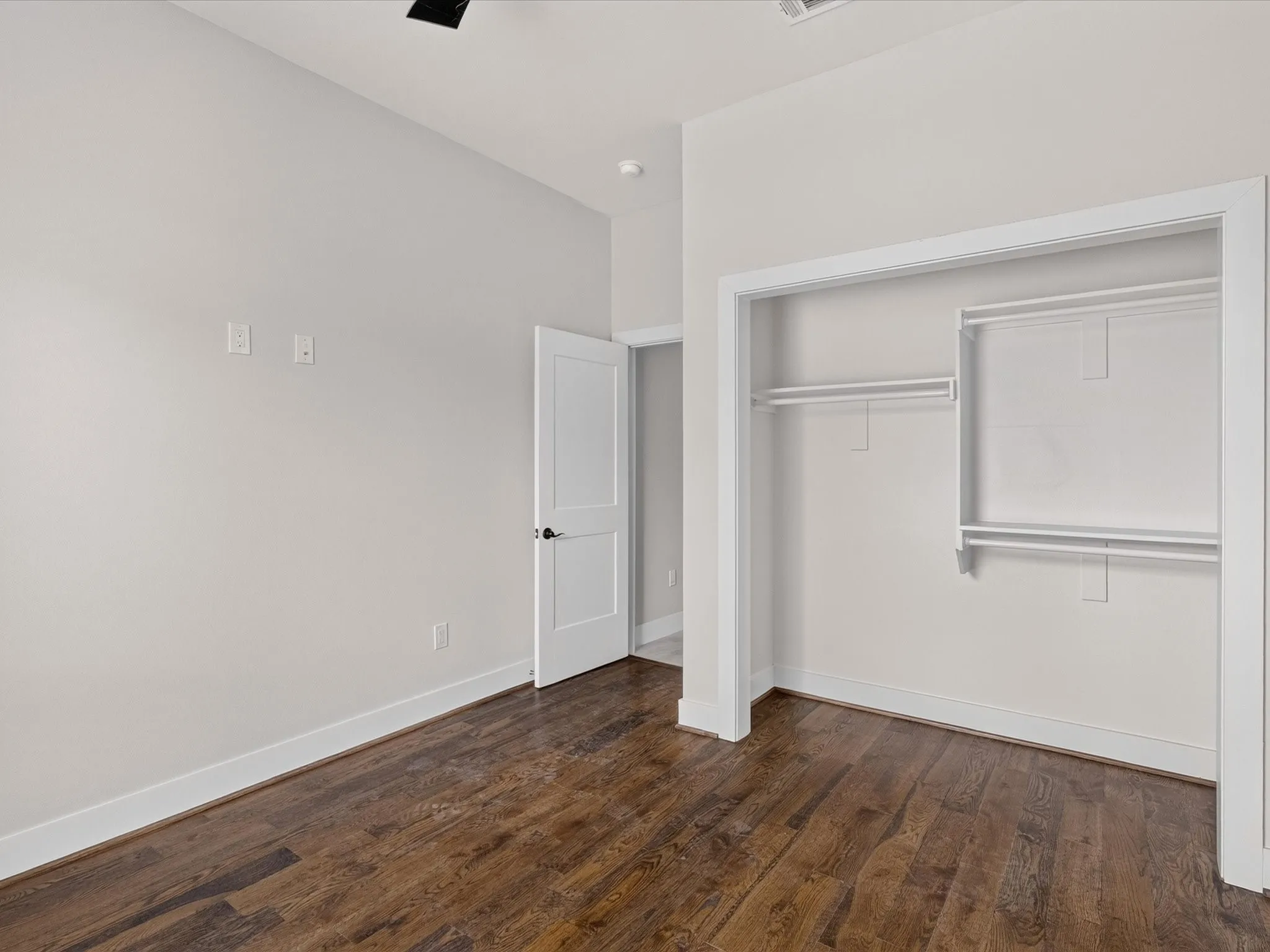 Wood-finish flooring, white trim, open closet with shelving and hanging rods, white interior door, and light-colored walls