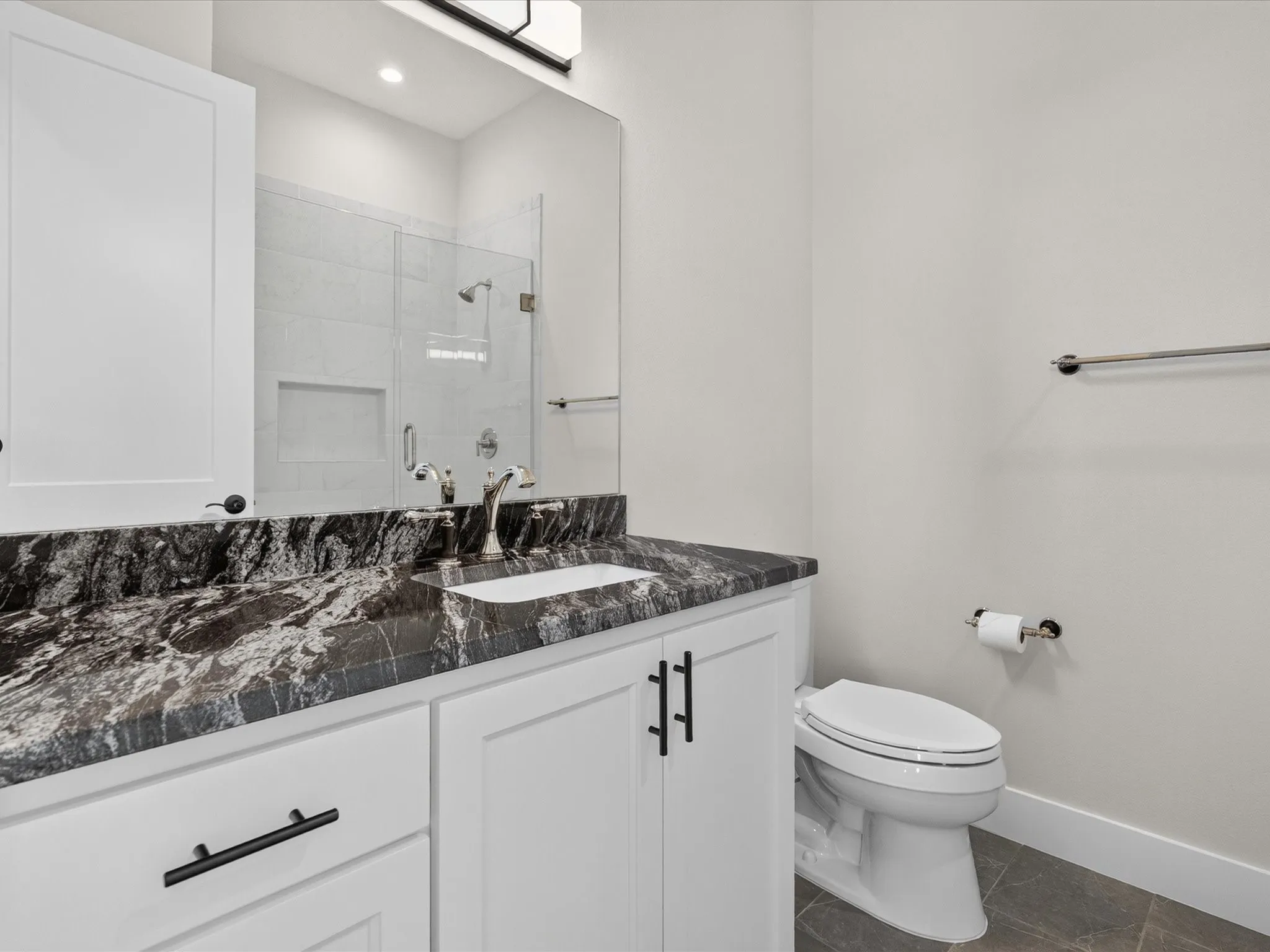 White shaker-style cabinetry with matte black hardware, dark patterned countertop with an undermount sink, frameless mirror, and a glass-enclosed shower with tile surround