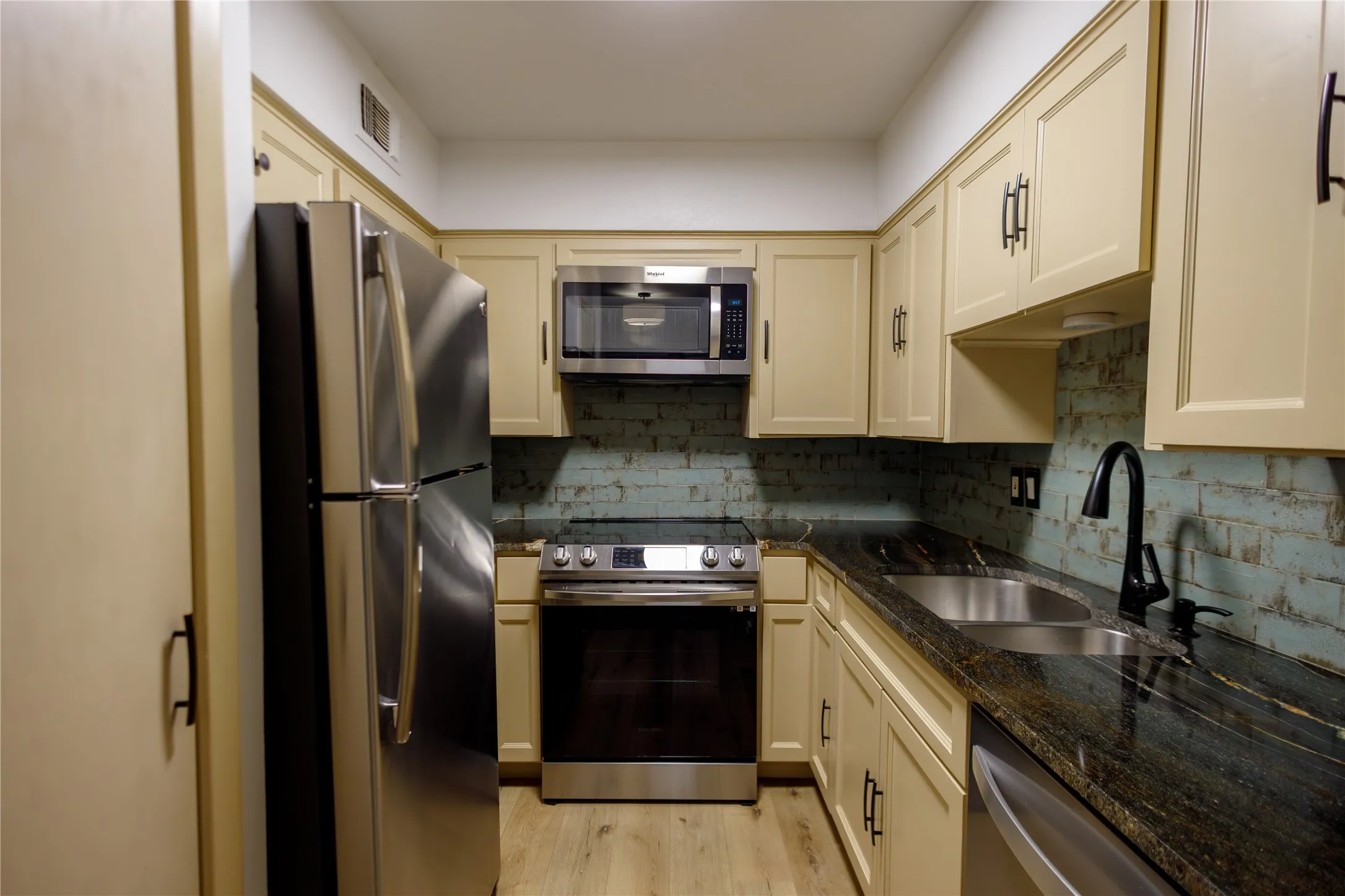 Kitchen featuring stainless steel appliances, dark stone countertops, light-toned cabinetry, and wood-finish flooring