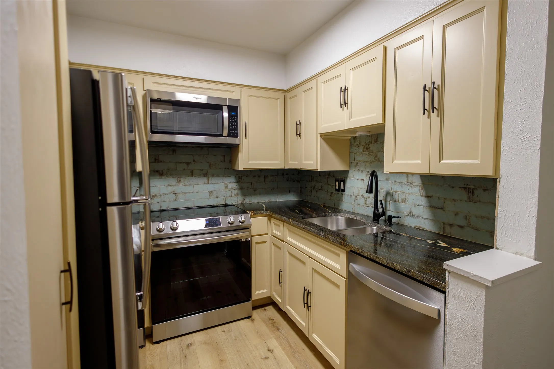 Kitchen featuring stainless steel appliances, light-toned cabinetry, dark countertops, a brick-patterned backsplash, and wood-finish flooring