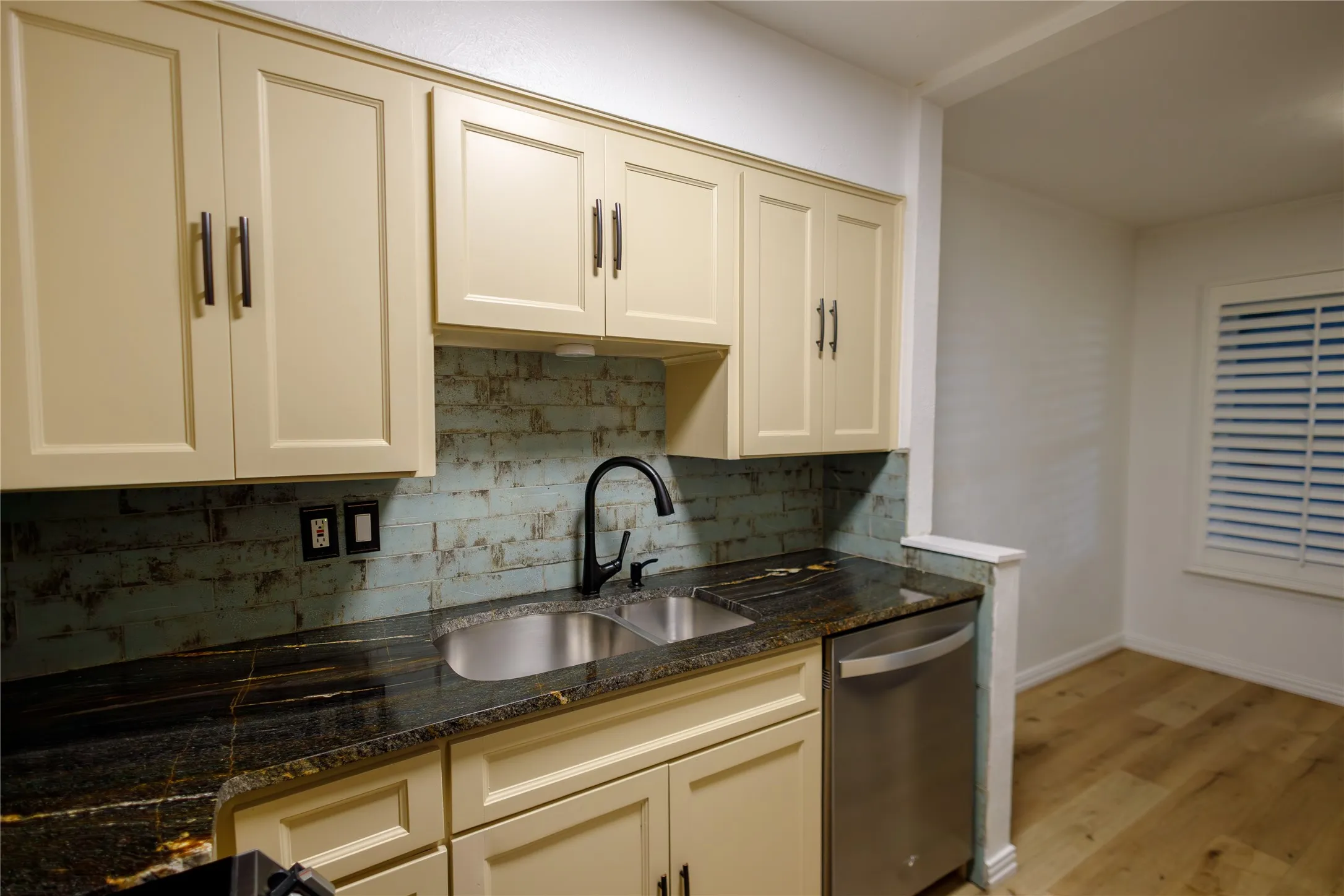 Cream-colored shaker-style cabinetry, dark stone countertops, stainless steel double basin sink, black gooseneck faucet, and a stainless steel dishwasher