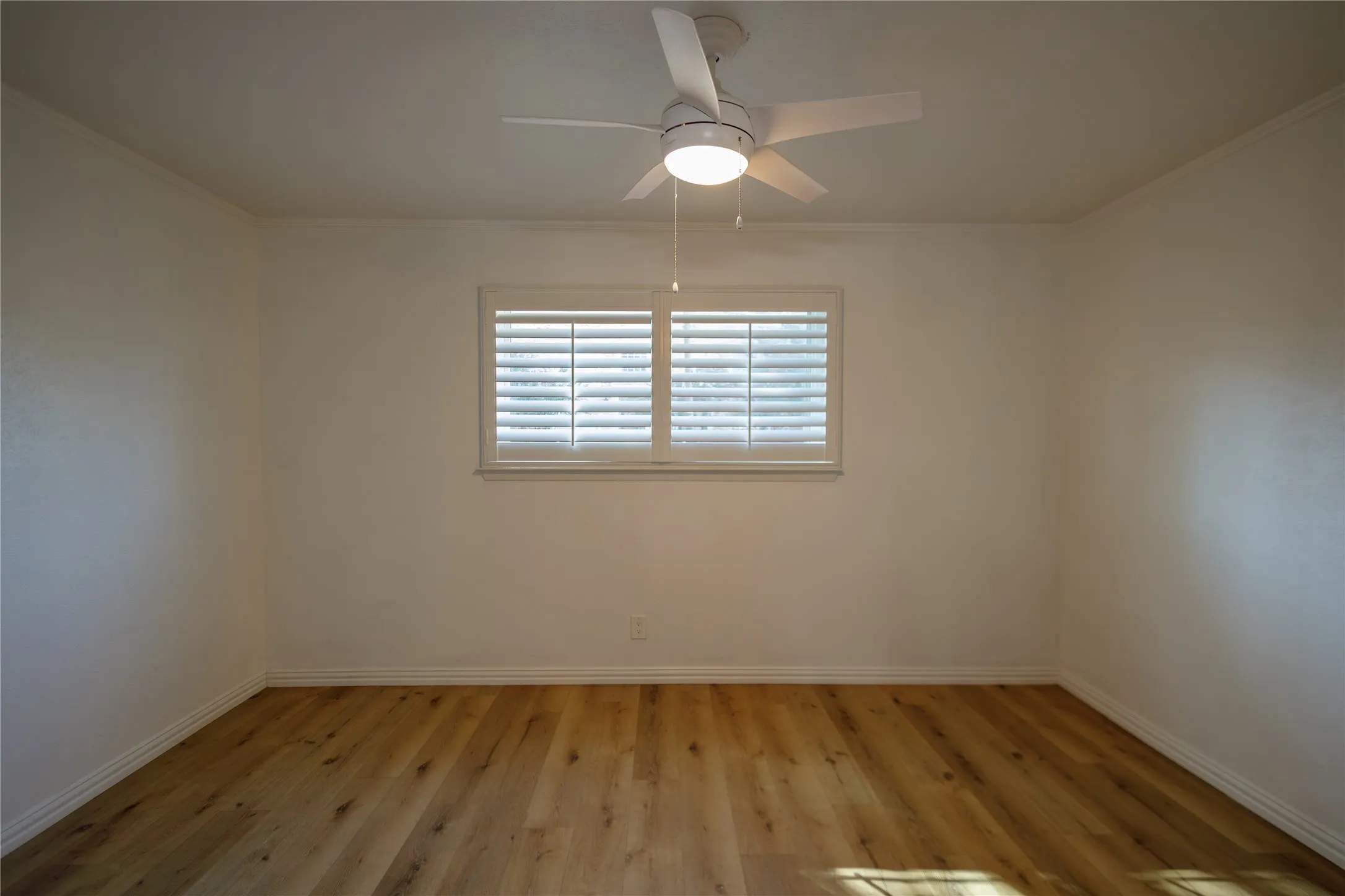 Wood-finish flooring, white walls, window with plantation shutters, and a ceiling fan with integrated lighting