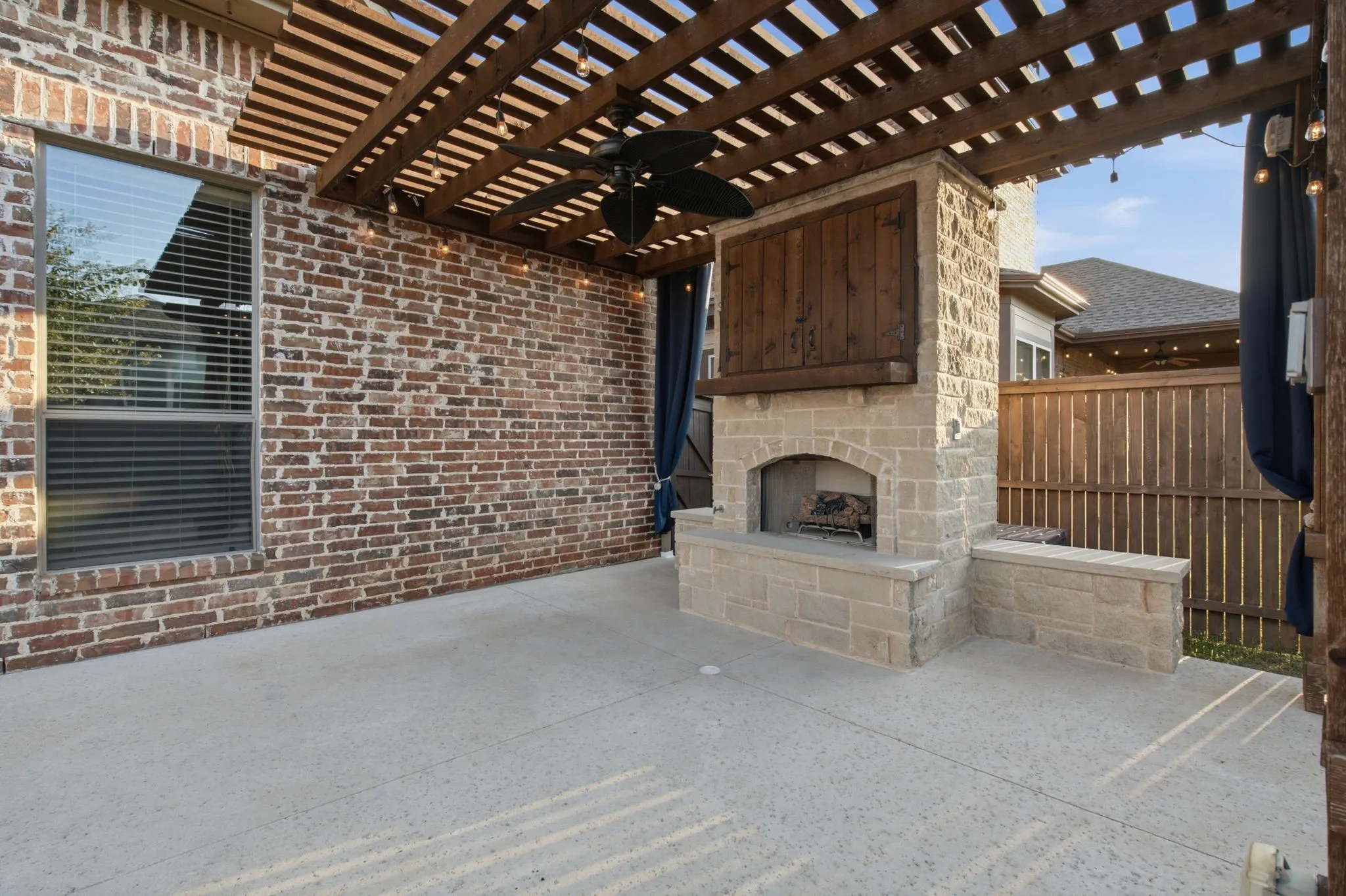 Expansive concrete patio featuring a wood pergola with integrated lighting, a ceiling fan, and a stone-clad outdoor fireplace with a wood mantle