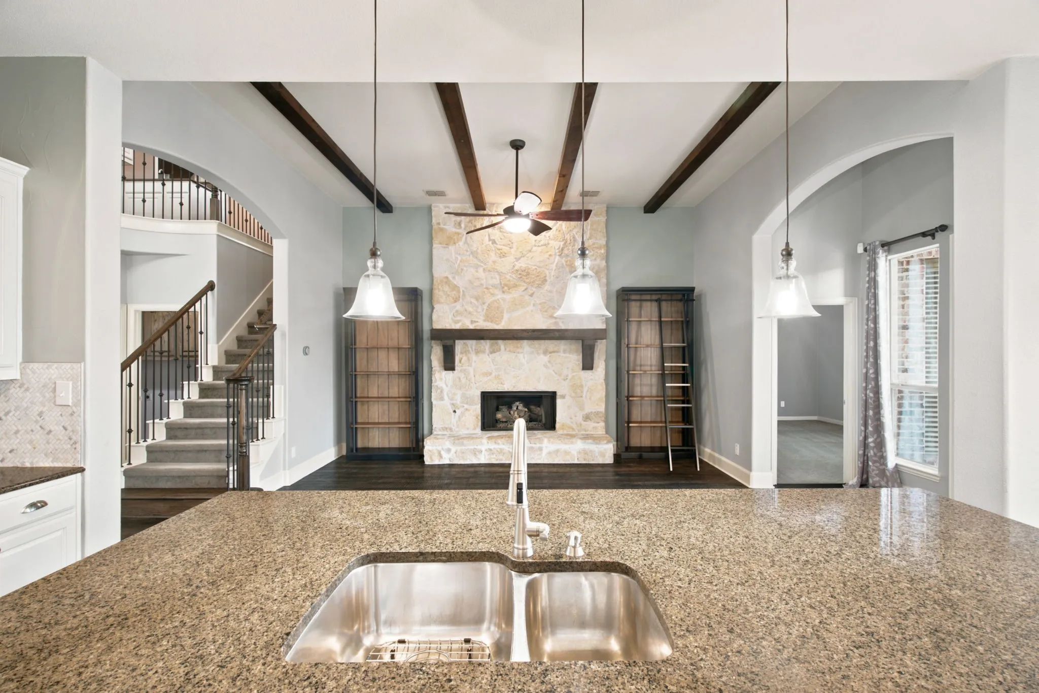 Kitchen island with a double stainless steel sink and granite countertop, with a stone fireplace featuring a wood mantel, exposed ceiling beams, and a ceiling fan in the living area