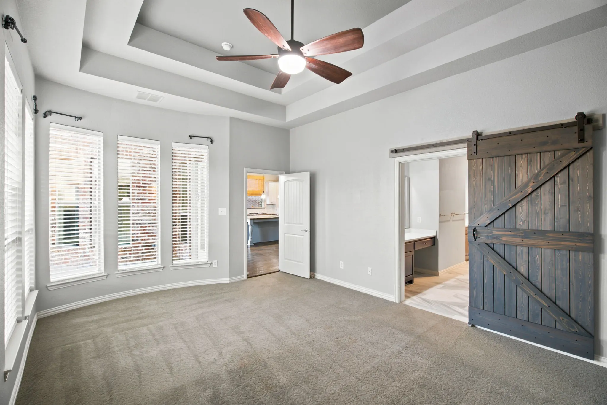 Tray ceiling with a ceiling fan, three vertical windows with blinds, and a dark wood-finish barn door