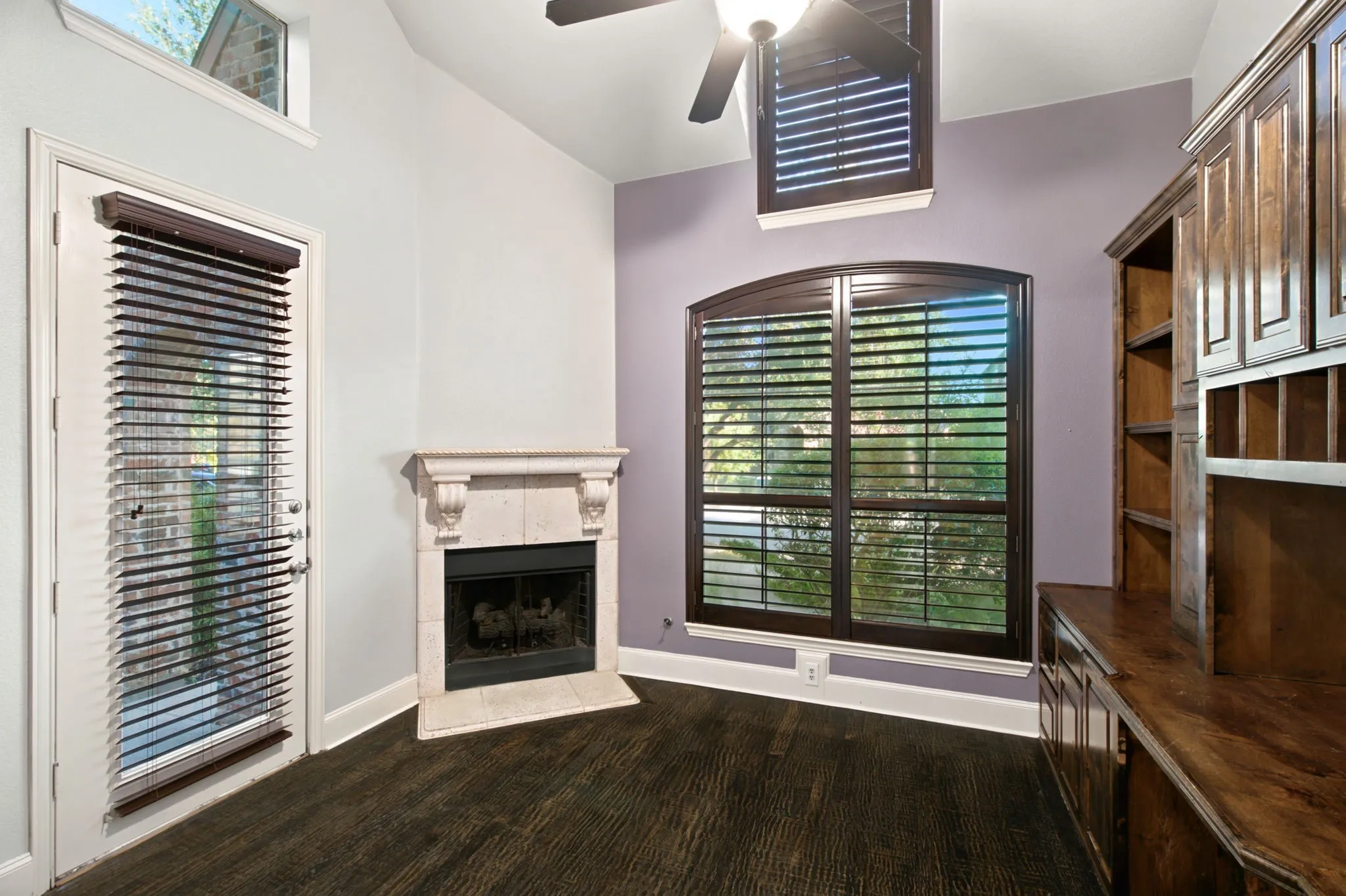 Imagine working with a fire to keep you warm in the winter months. Dark wood-finish flooring, built-in wooden cabinetry, fireplace with a stone mantel, arched window with plantation shutters, and a ceiling fan