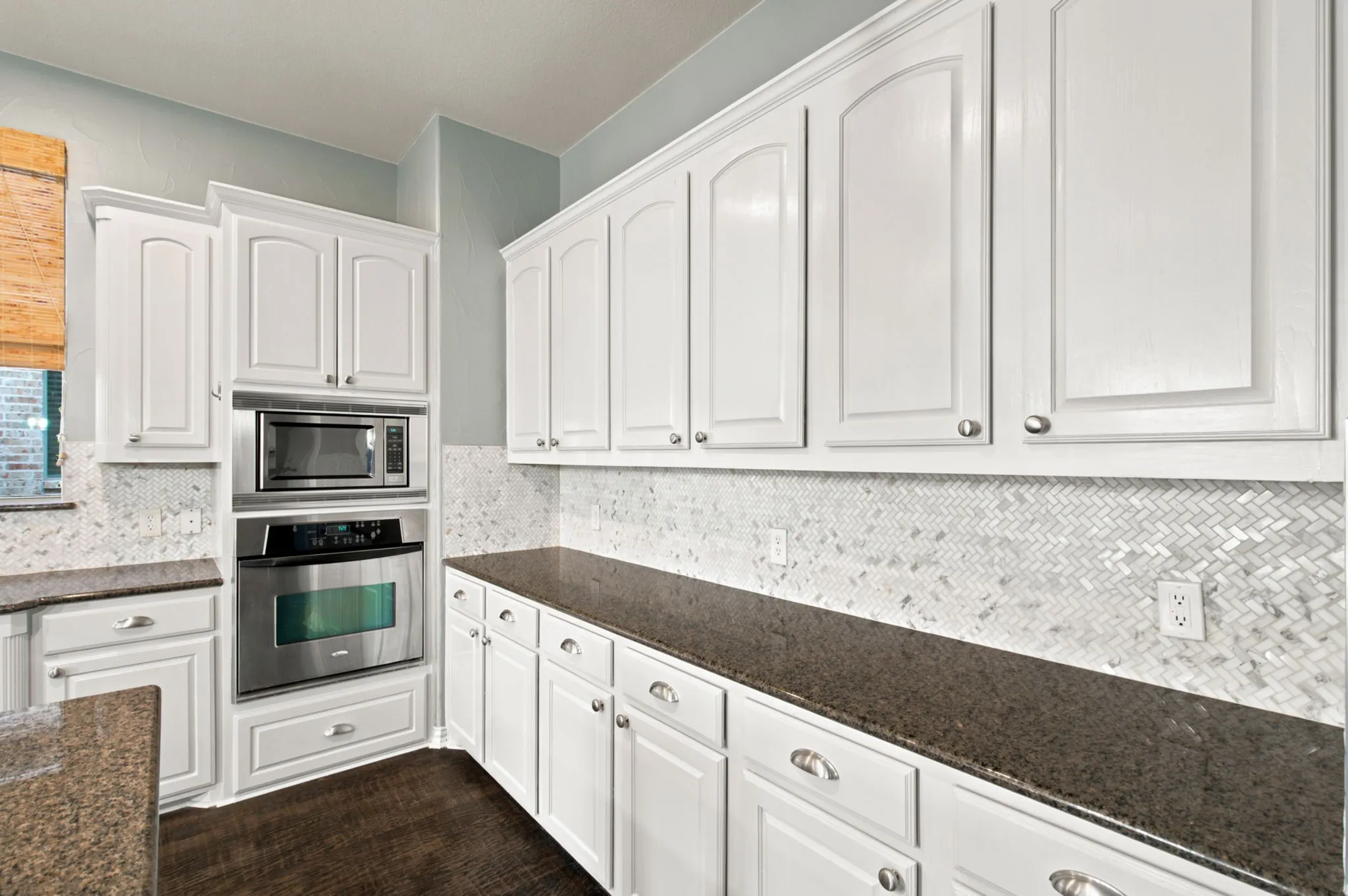 White cabinetry with brushed nickel hardware, dark stone countertops, herringbone-patterned tile backsplash, and built-in stainless steel microwave and oven