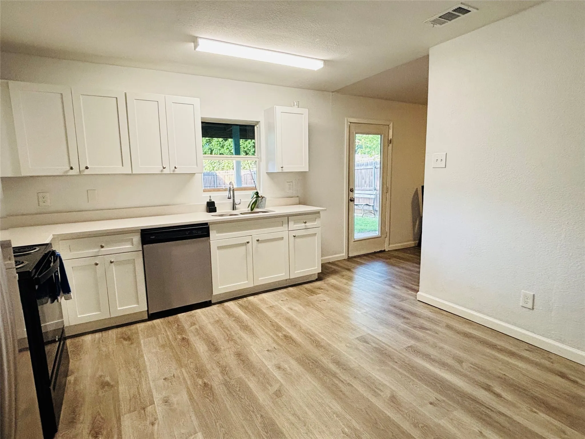 White shaker cabinetry, light-toned countertops, stainless steel dishwasher, wood-finish flooring, and a glass-paneled door