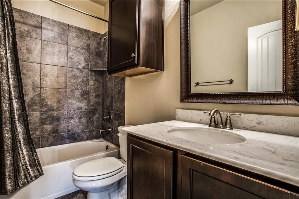 Bathroom featuring a white porcelain tub, dark-toned tile surround, dark wood-finish vanity with a single sink, light-toned countertop, and framed mirror