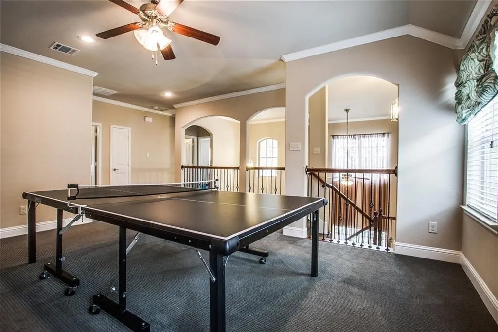 Carpeted loft area featuring a ceiling fan, crown molding, arched entryways, and a window with blinds
