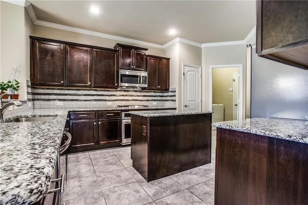 Kitchen featuring granite countertops, wood-finish cabinetry, tile flooring, stainless steel appliances, and a tile backsplash