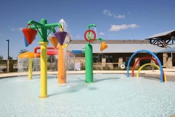 Outdoor splash pad with multiple colorful water features, including tipping buckets and arch sprays, complemented by a nearby stone-clad building with a metal roof