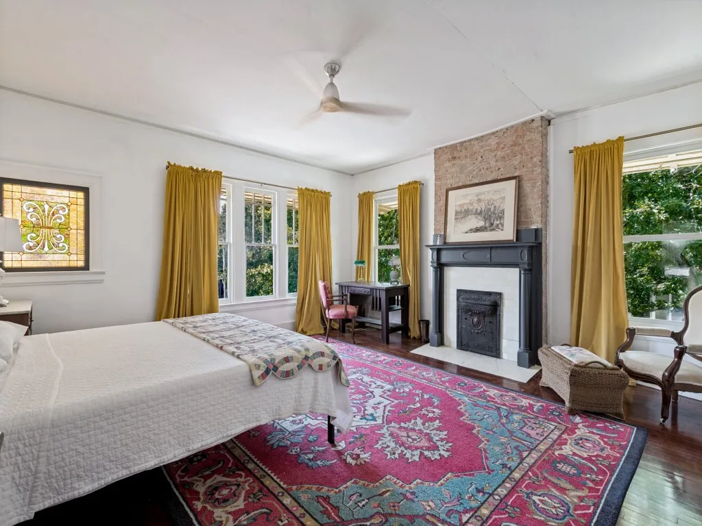 Spacious primary bedroom featuring hardwood flooring, a fireplace with black surround and mantelpiece, ceiling fan, and multiple tall windows dressed in gold drapery