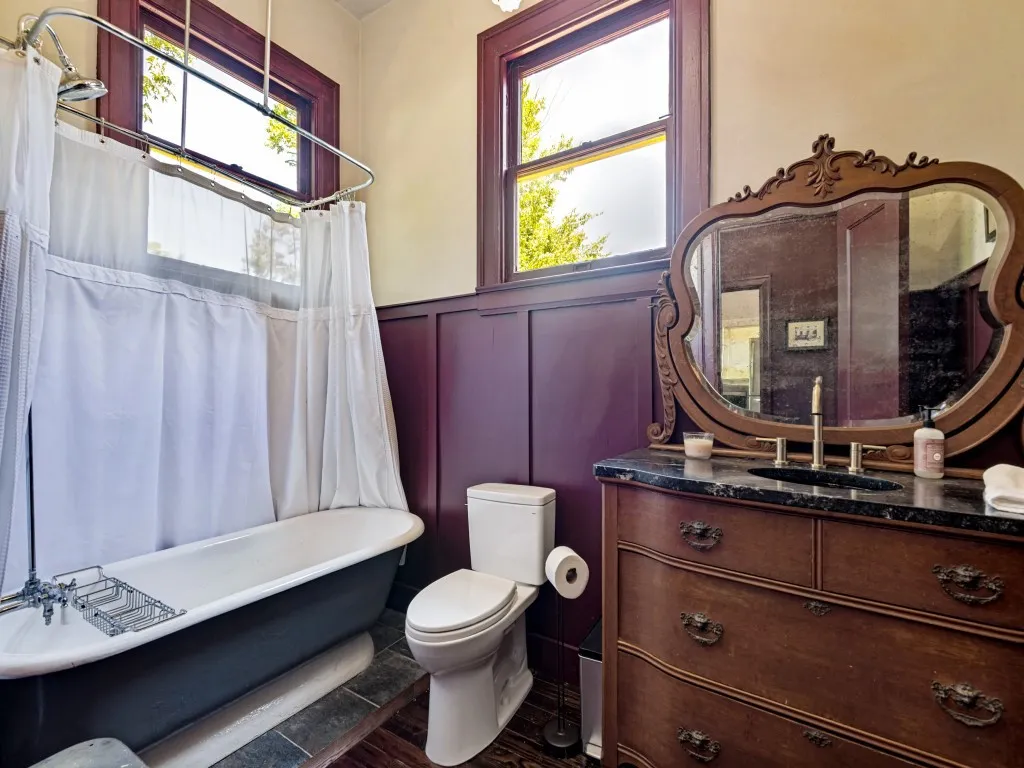 Bathroom featuring a freestanding pedestal  tub with shower, rich wood paneling, dark stone vanity countertop, an ornate wood-framed mirror, and wood-finish flooring