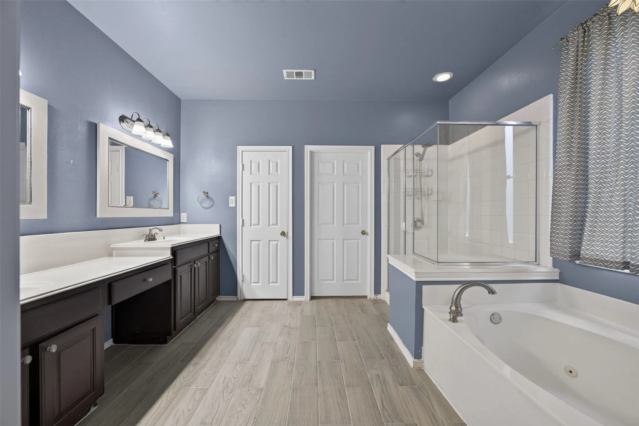 Dual vanity with white countertops, wood-finish flooring, built-in soaking tub, and enclosed glass shower