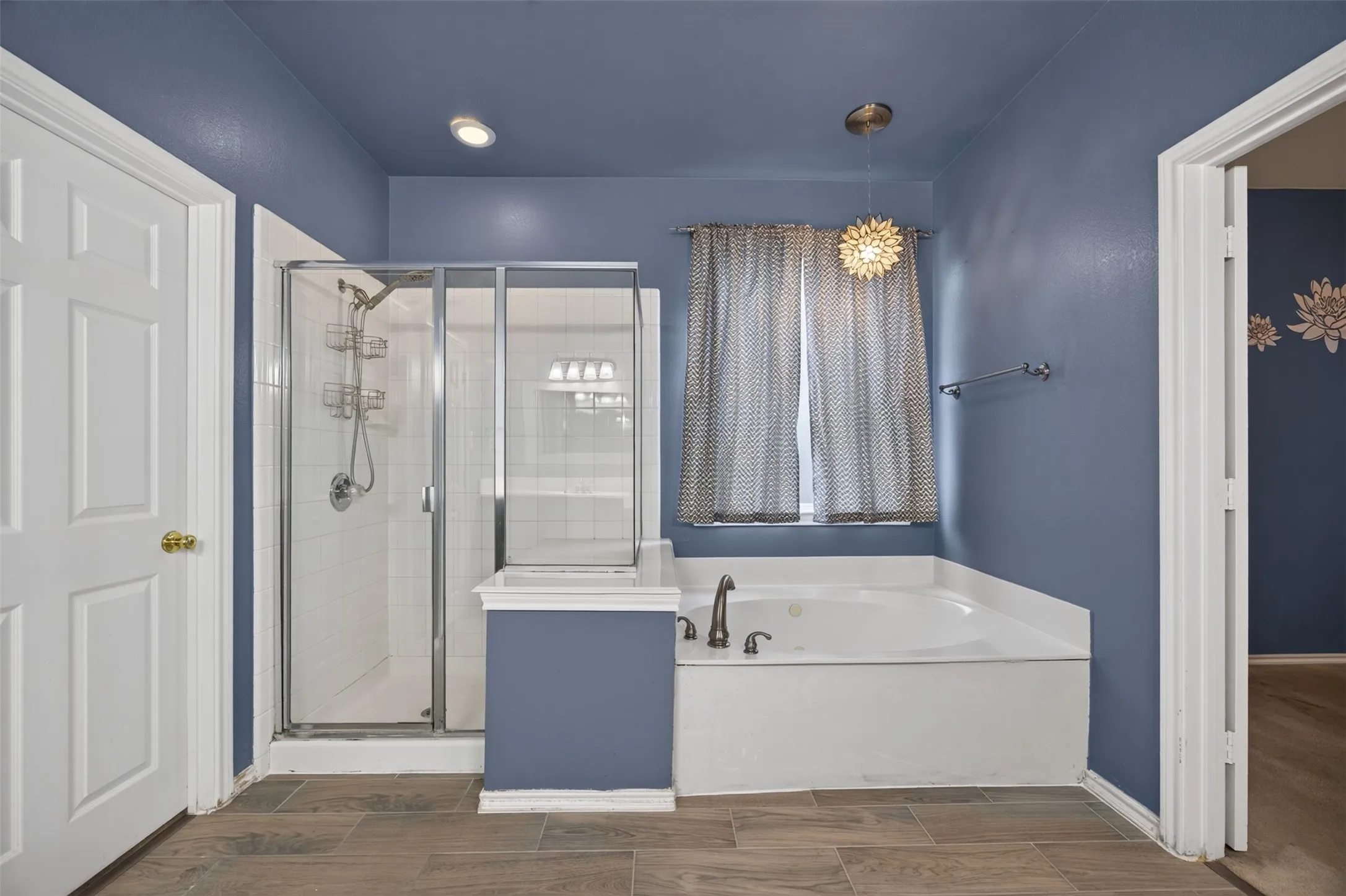 Spacious bath featuring a built-in soaking tub, a separate tiled shower with glass enclosure, wood-finish flooring, and a decorative pendant light fixture