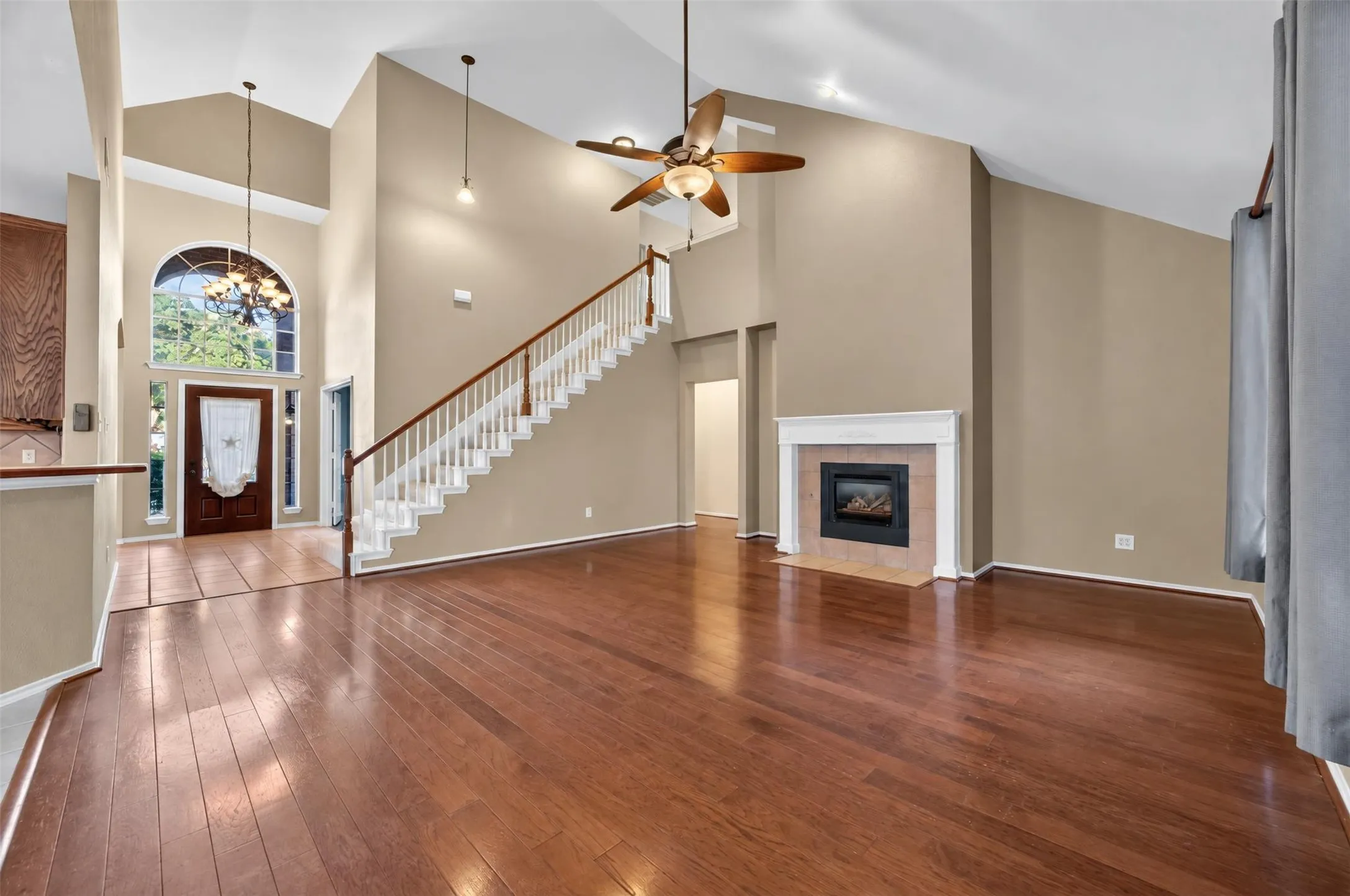 Vaulted ceiling with an overhead fan, wood-finish flooring, staircase with white risers and a wood handrail, fireplace with a white mantel, and an arched transom window above the entry