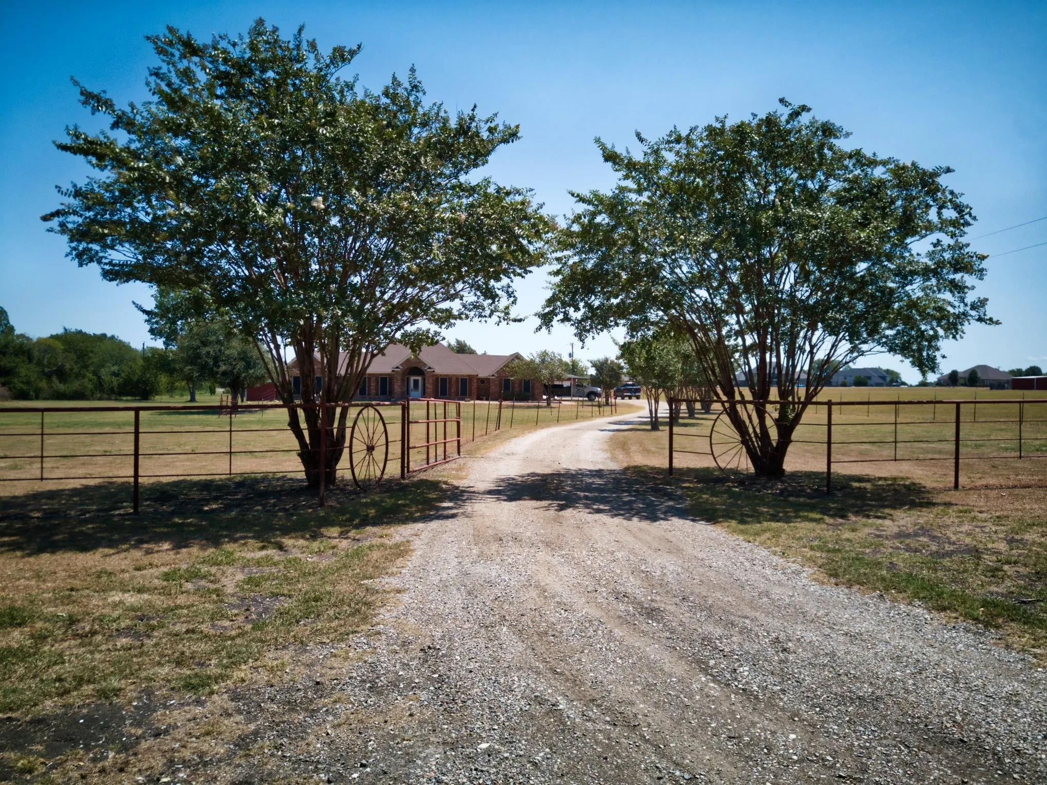 Gravel driveway entry featuring mature trees and a metal perimeter fence
