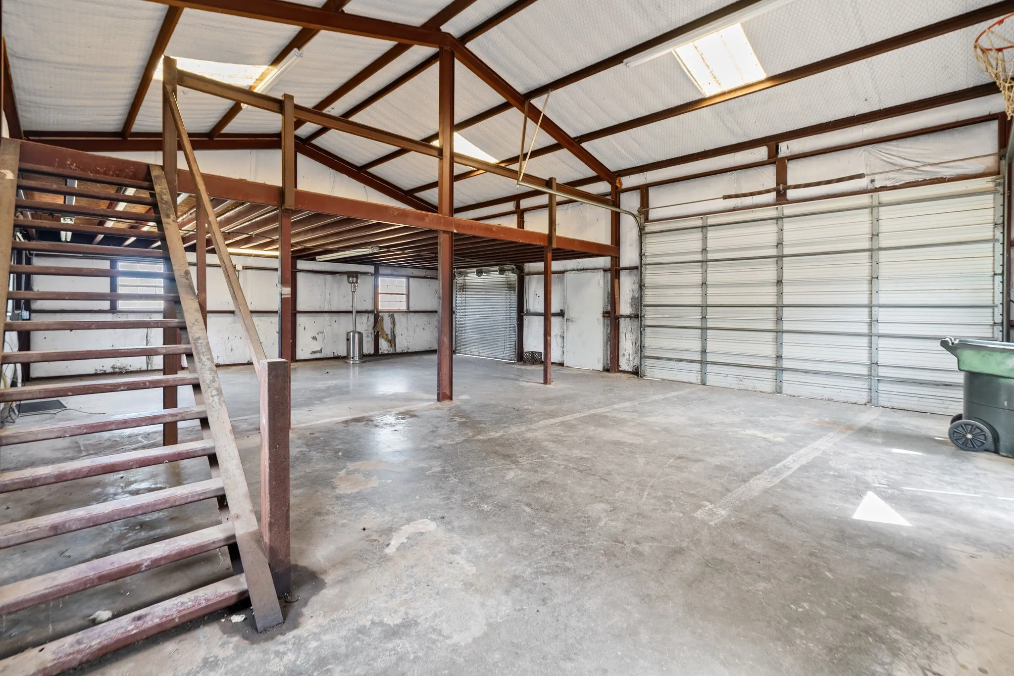 Large open space with concrete flooring, exposed metal beam structure, upper level mezzanine, industrial roll-up door, and a skylight