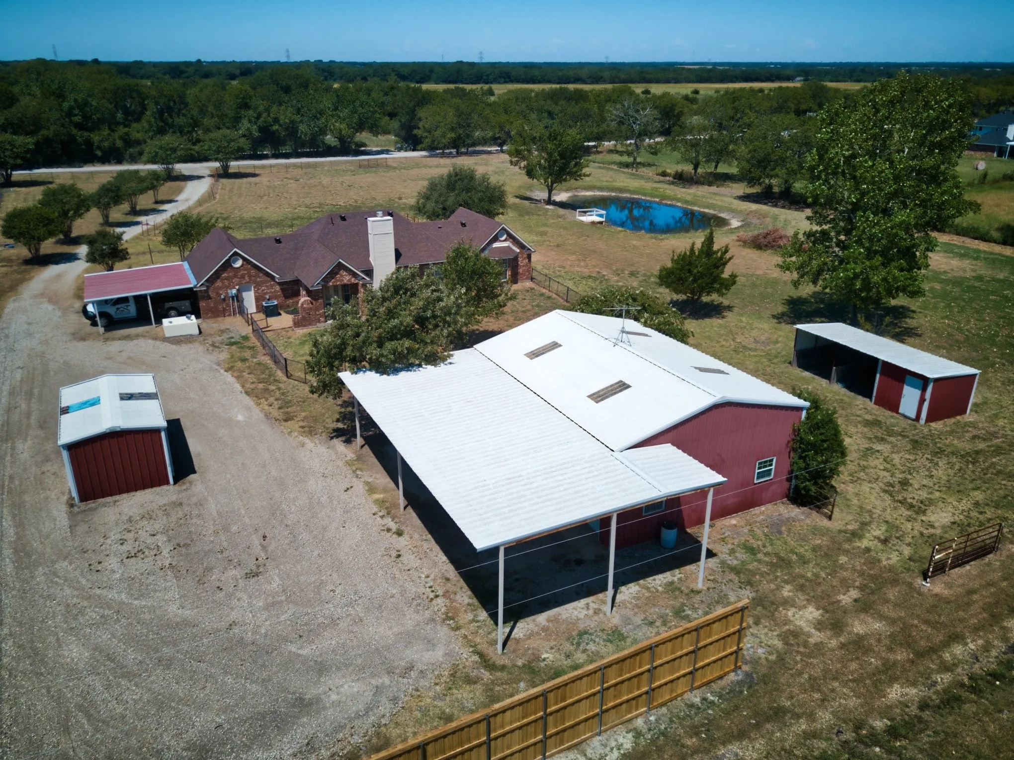 Red brick house with a brown shingle roof, accompanied by a detached red barn with a white metal roof and multiple skylights, a red storage shed, a covered carport, and a pond