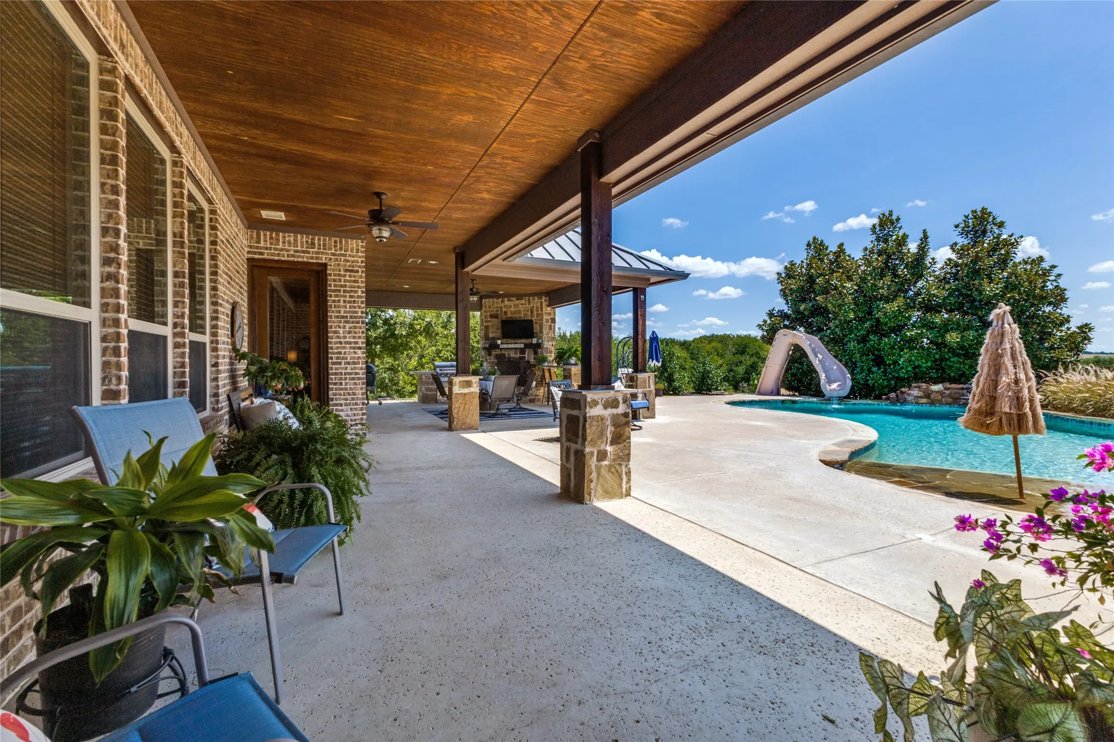 Expansive covered patio with exposed wood-finish ceiling, brick facade, stone columns, and a built-in outdoor kitchen