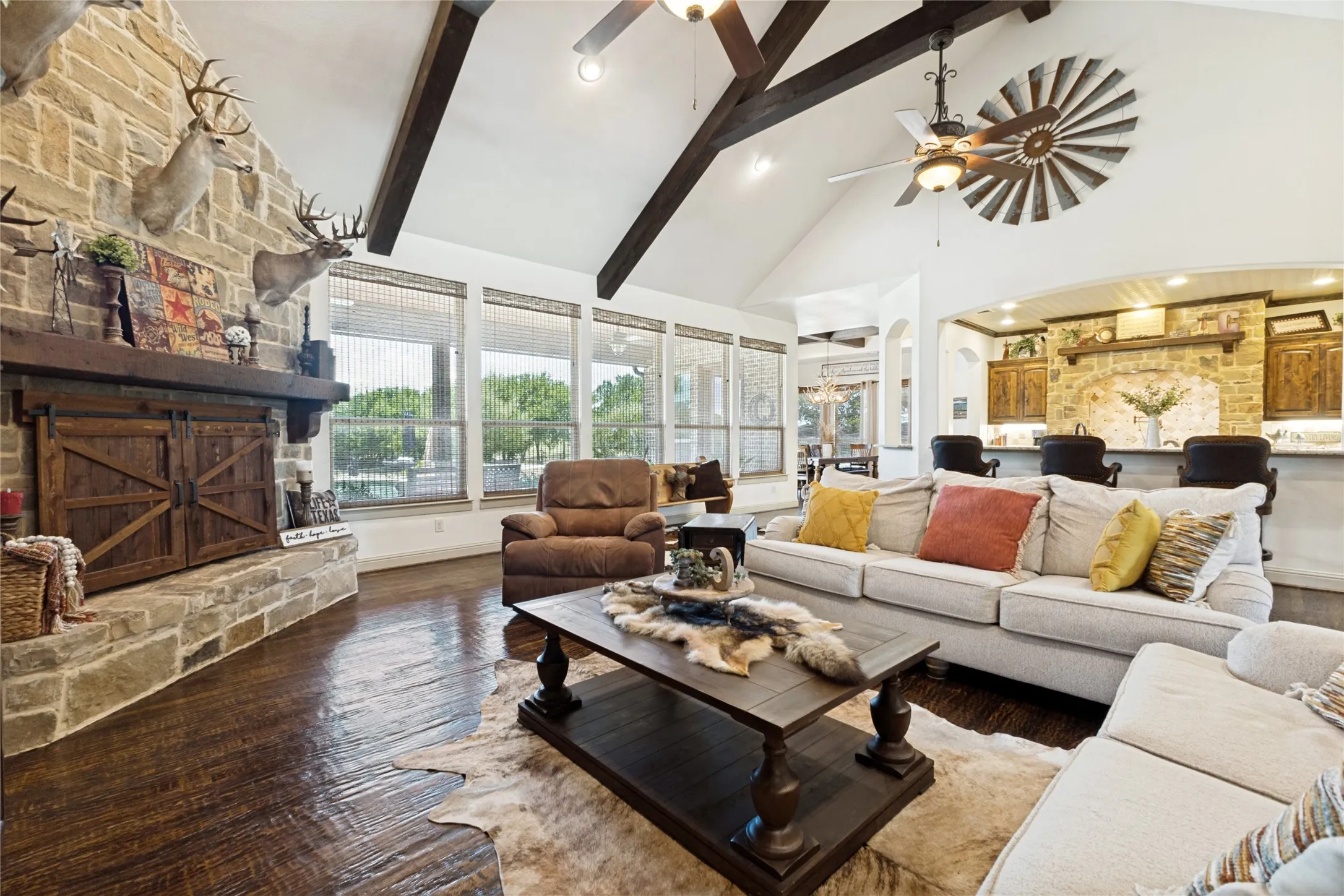 Stone-clad fireplace with a wood mantle, vaulted ceiling with exposed wood beams, wood-finish flooring, and a kitchen bar with stone detailing