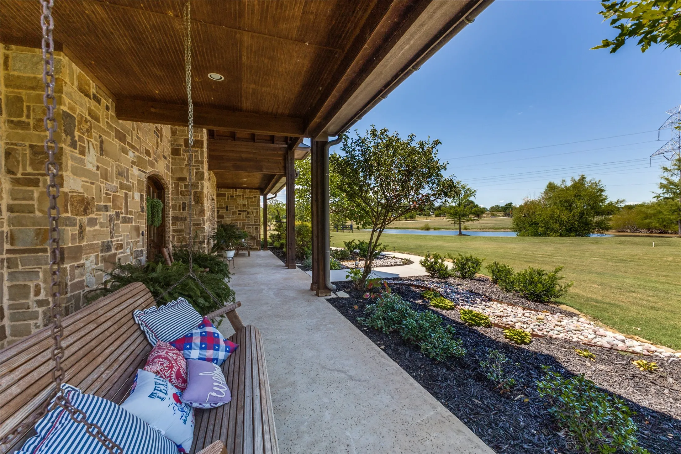 Covered stone-clad porch with wood-paneled ceiling, concrete flooring, and manicured landscaping featuring a water feature
