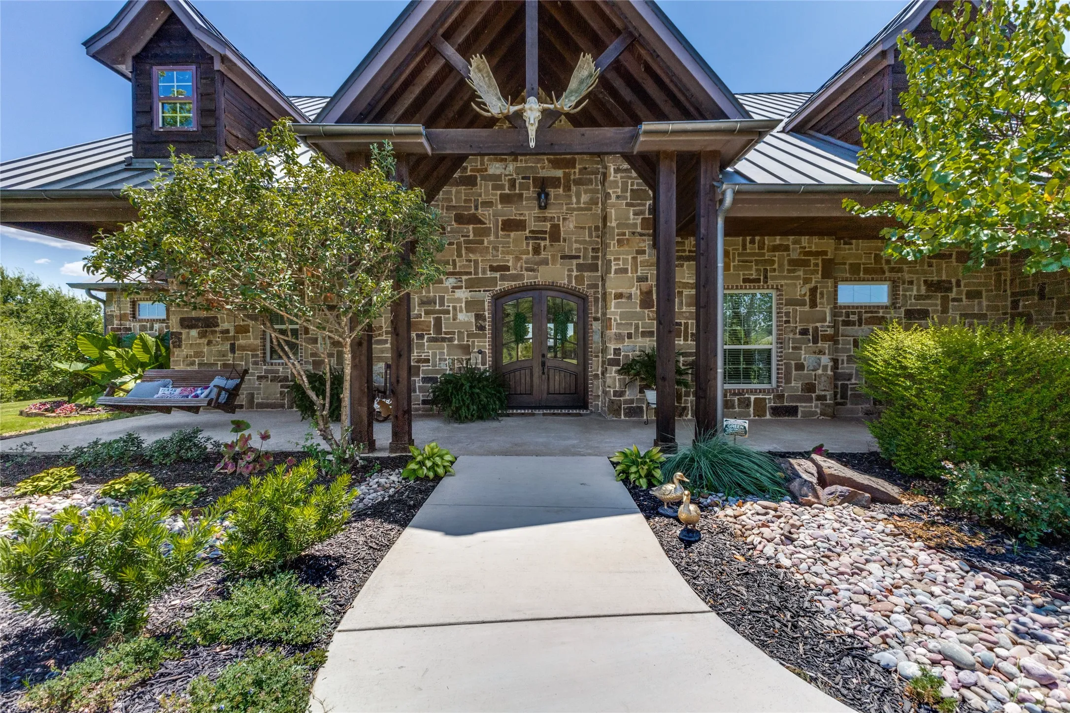 Stone exterior with a metal roof, featuring a covered entry porch with timber beams, double arched entry doors, and a concrete walkway