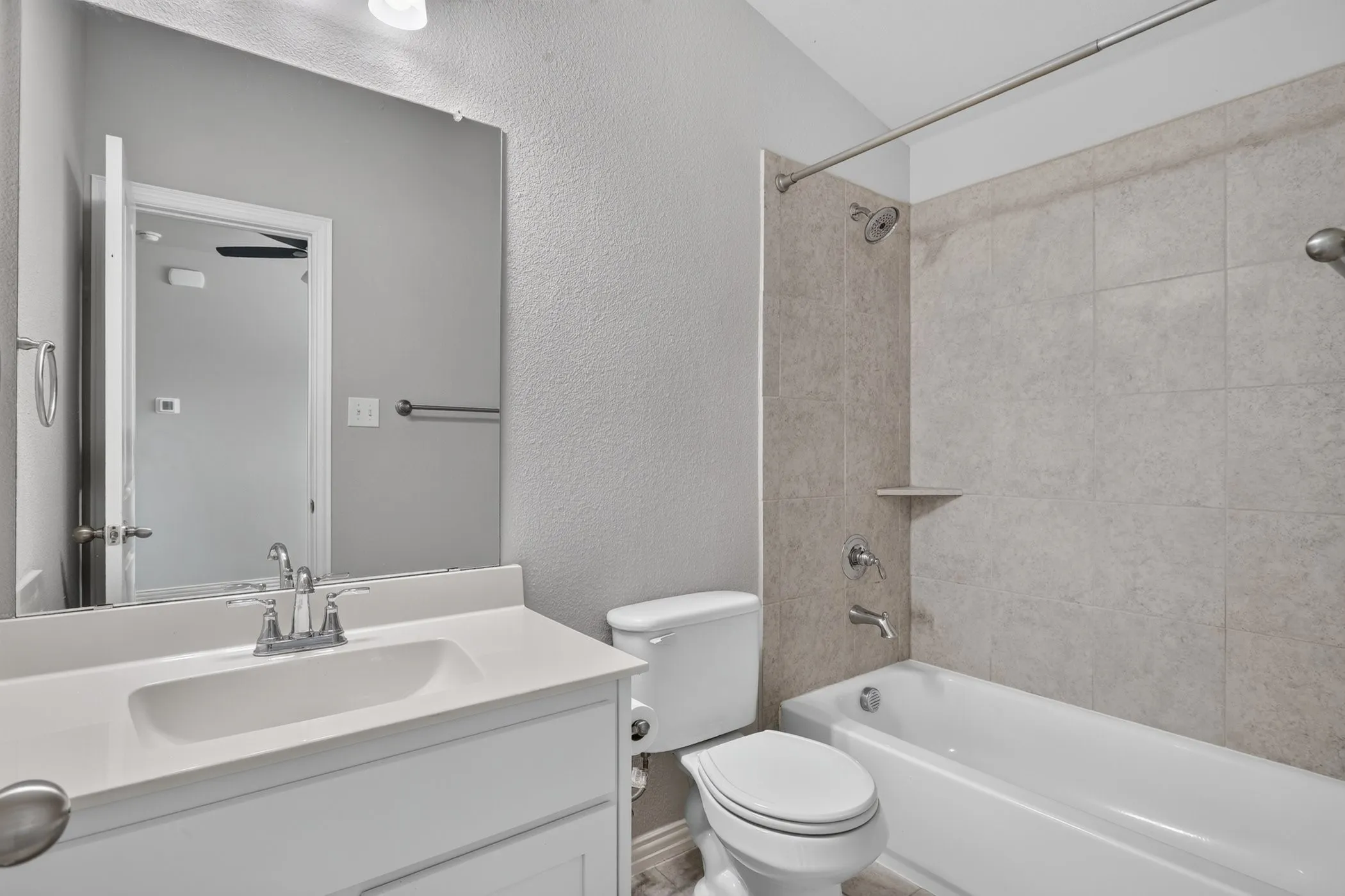 White vanity with integrated sink and chrome faucet, large wall mirror, white bathtub, tiled shower surround with built-in shelf, and a white toilet