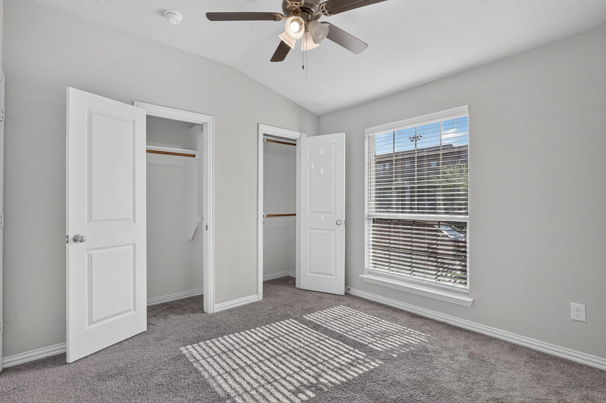 Vaulted ceiling with ceiling fan, two spacious closets with built-in shelving, plush carpeting, and a large window with horizontal blinds