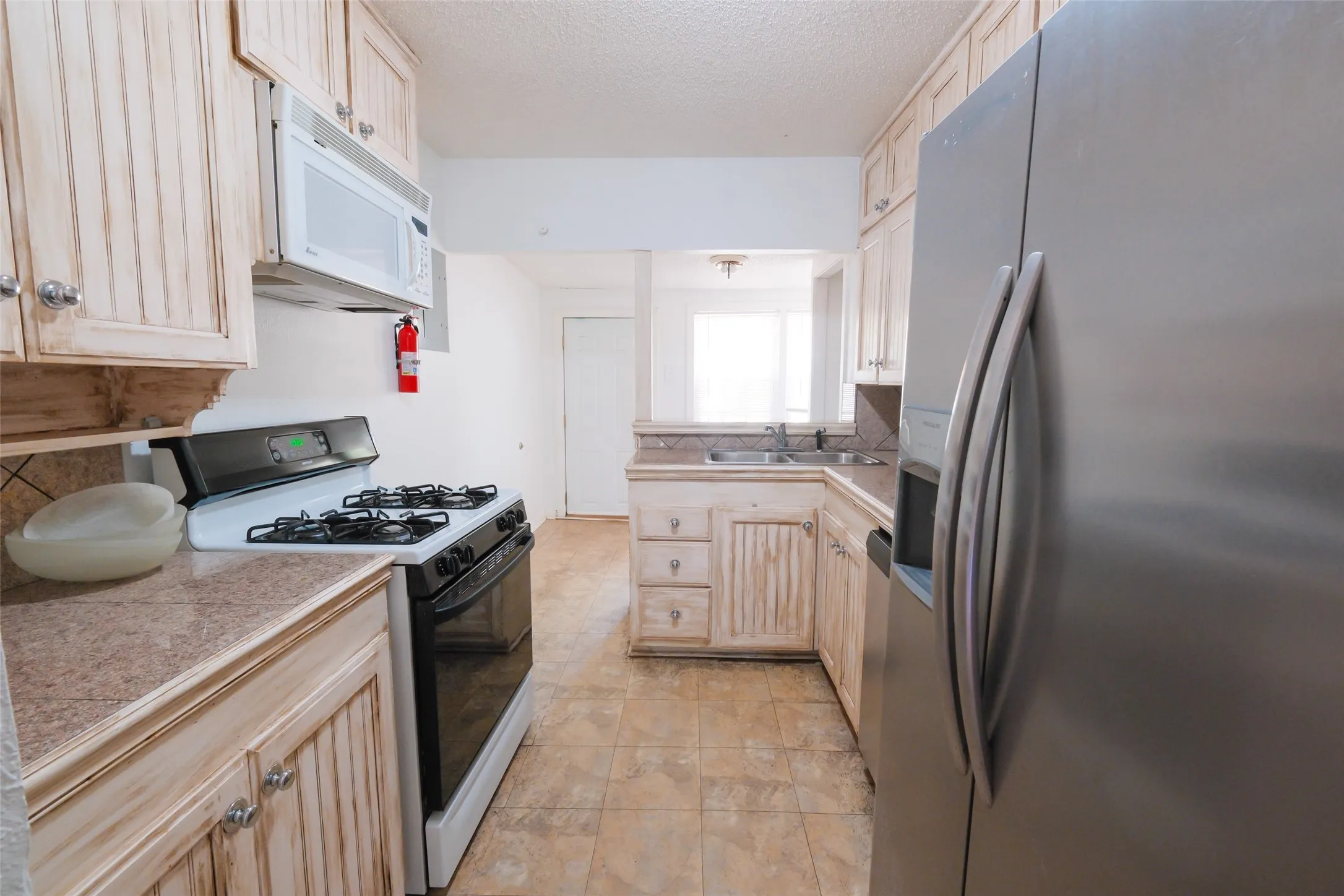 Galley kitchen featuring light-toned cabinetry, a stainless steel refrigerator, white gas range with oven, and a built-in microwave