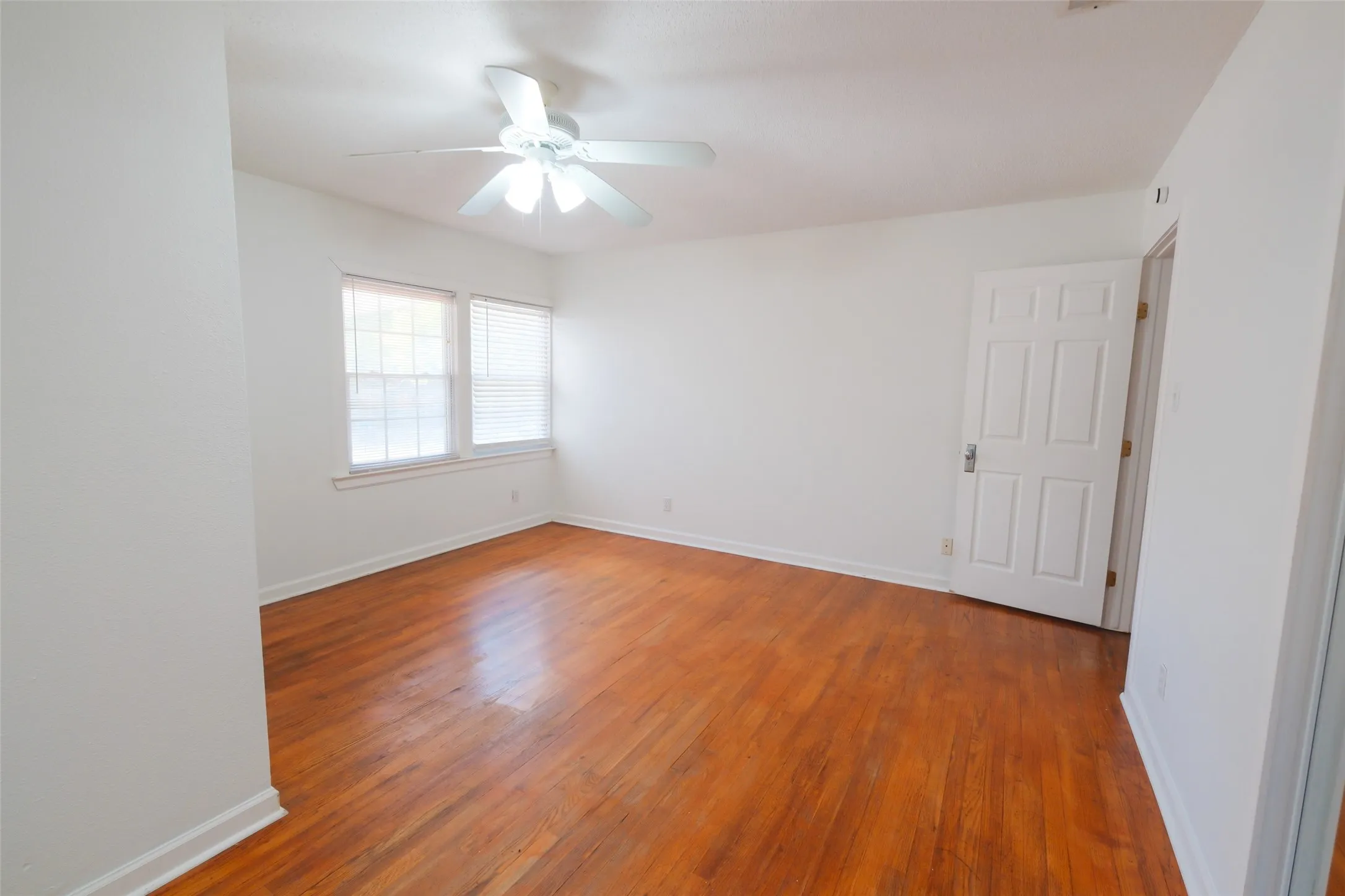 Hardwood flooring, white walls, two windows with blinds, ceiling fan with integrated lighting, and a paneled door