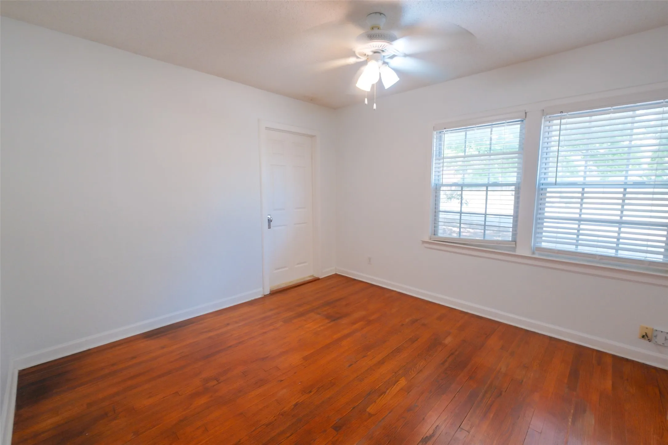 Hardwood flooring, white walls, two windows with blinds, ceiling fan, and a paneled door