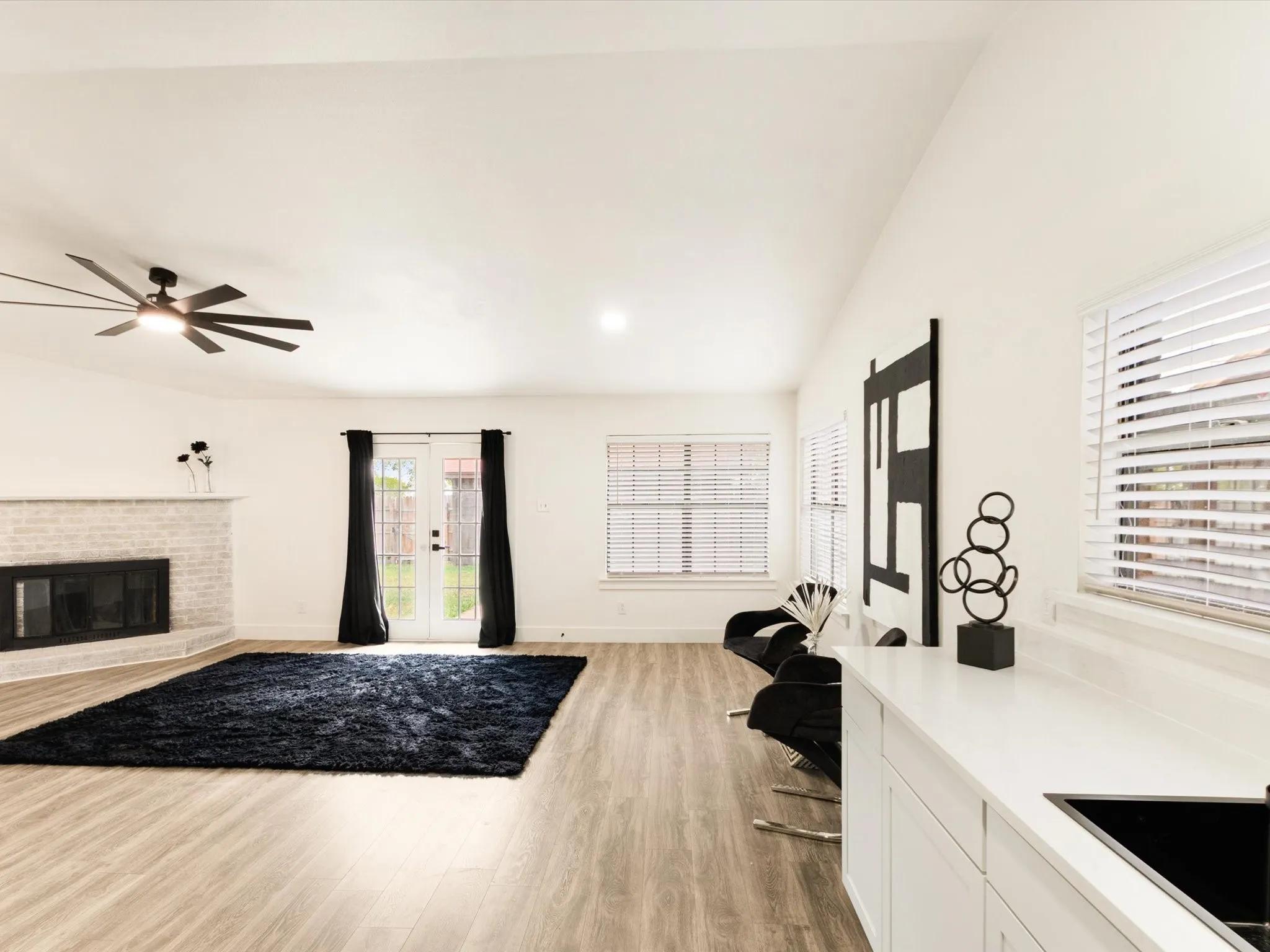 Vaulted ceiling with a ceiling fan, white brick fireplace, wood-finish flooring, white cabinetry with a built-in sink, and white window blinds