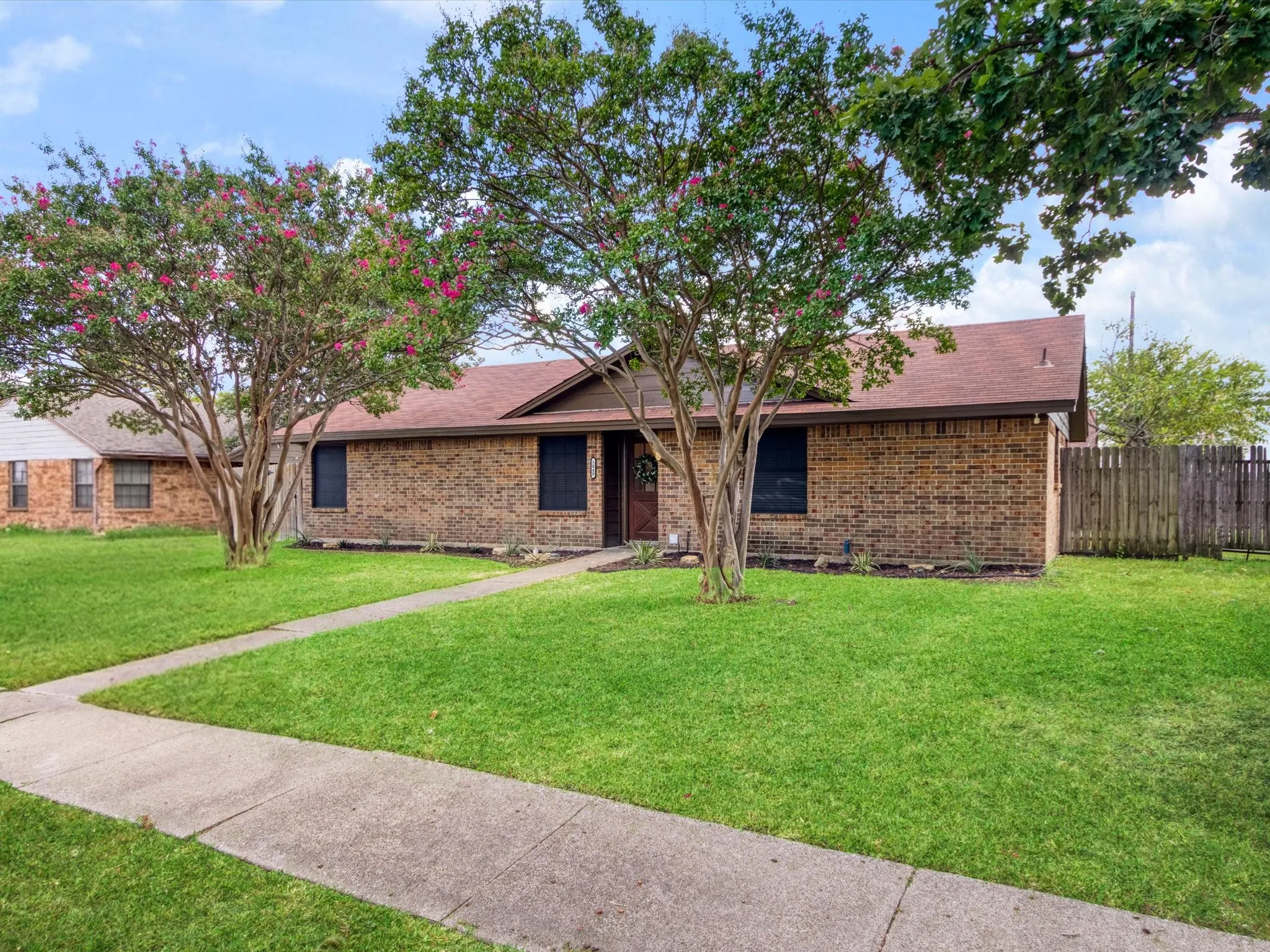 Brick exterior with a brown shingle roof, mature flowering trees, a concrete walkway, and a private wooden fence