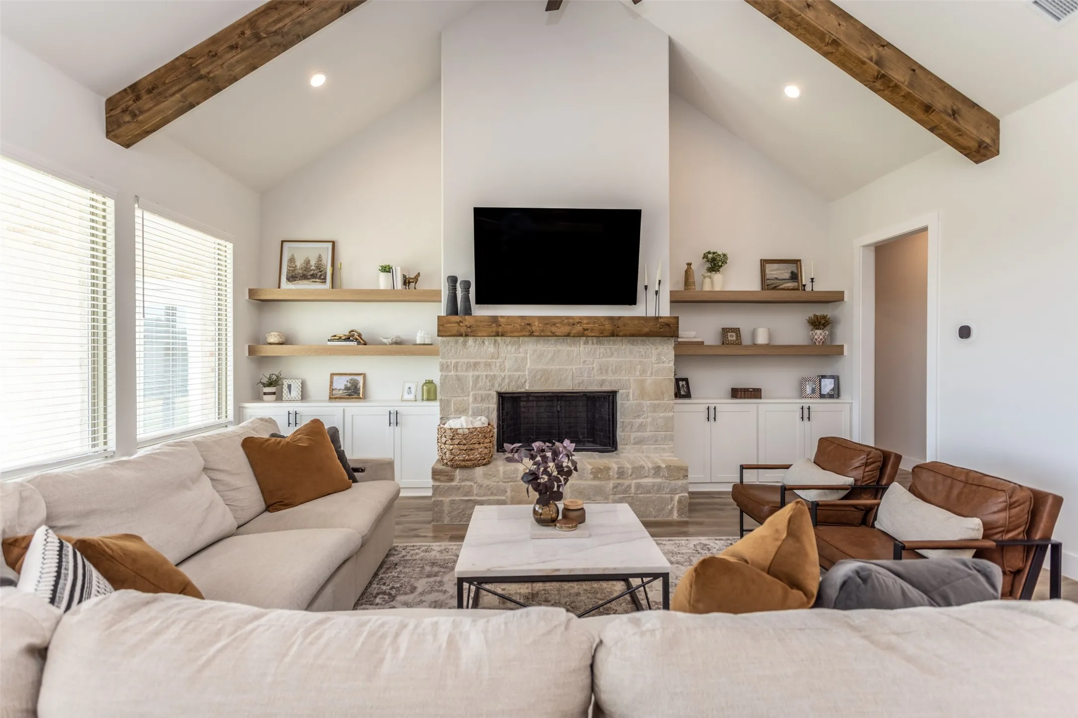 Vaulted ceilings with exposed wood beams, stone-clad fireplace, integrated built-in cabinetry, floating wood shelving, and wood-finish flooring
